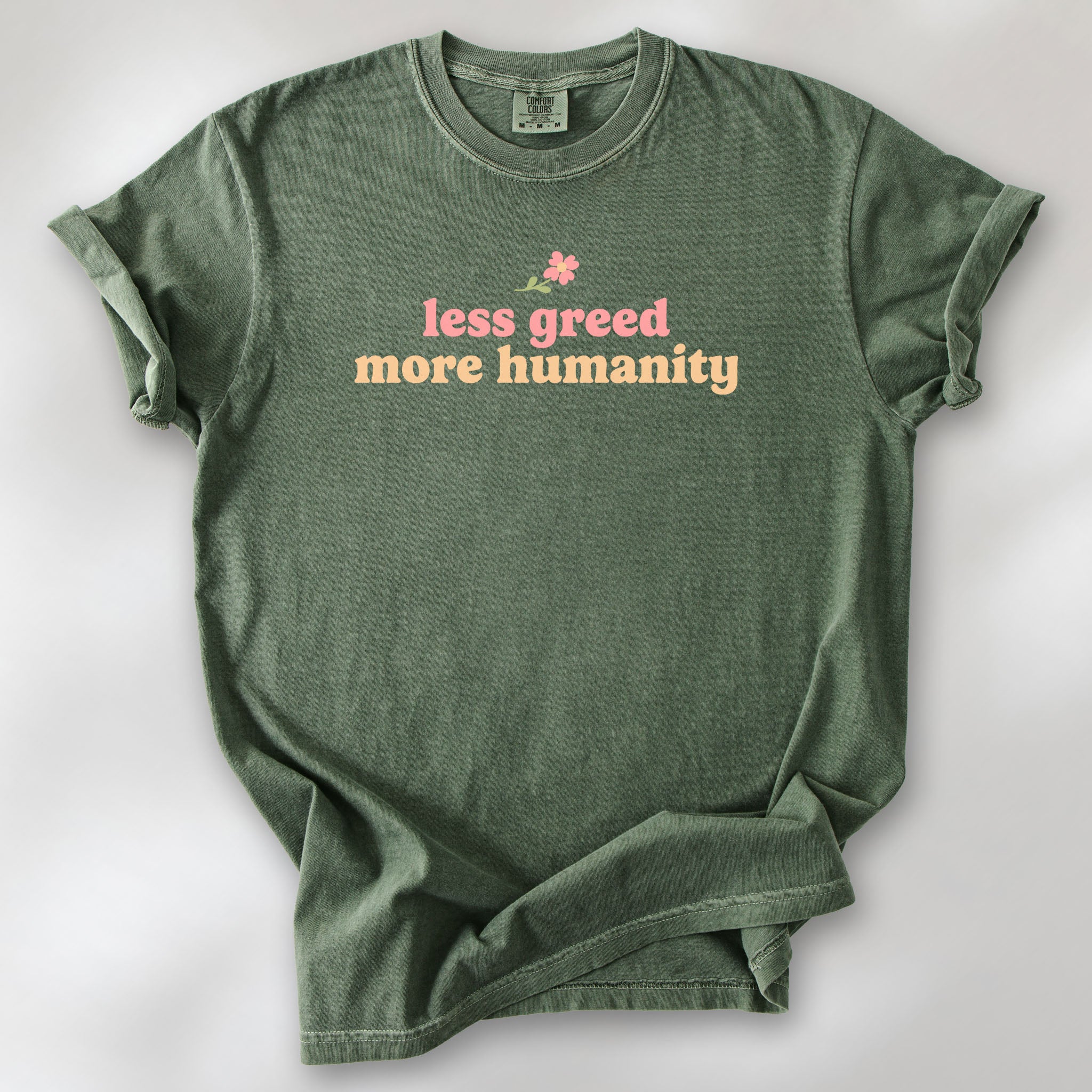 Moss t-shirt featuring the phrase "Less Greed More Humanity" in soft retro typography with a small flower graphic above the text.