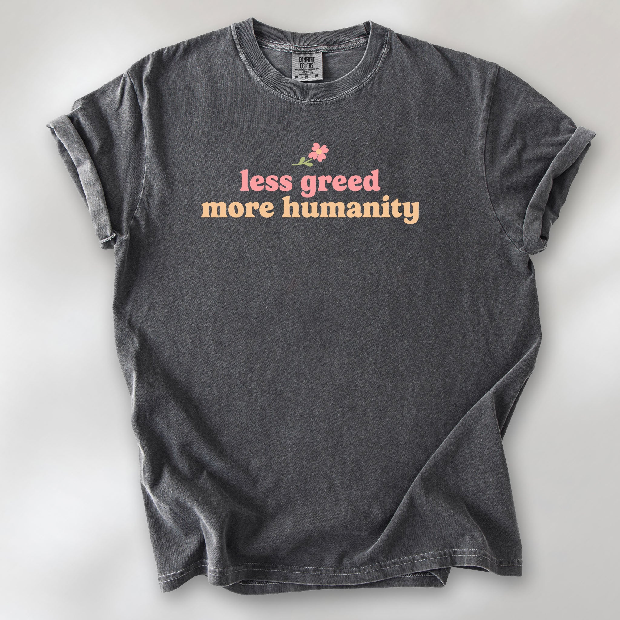 Pepper t-shirt featuring the phrase "Less Greed More Humanity" in soft retro typography with a small flower graphic above the text.