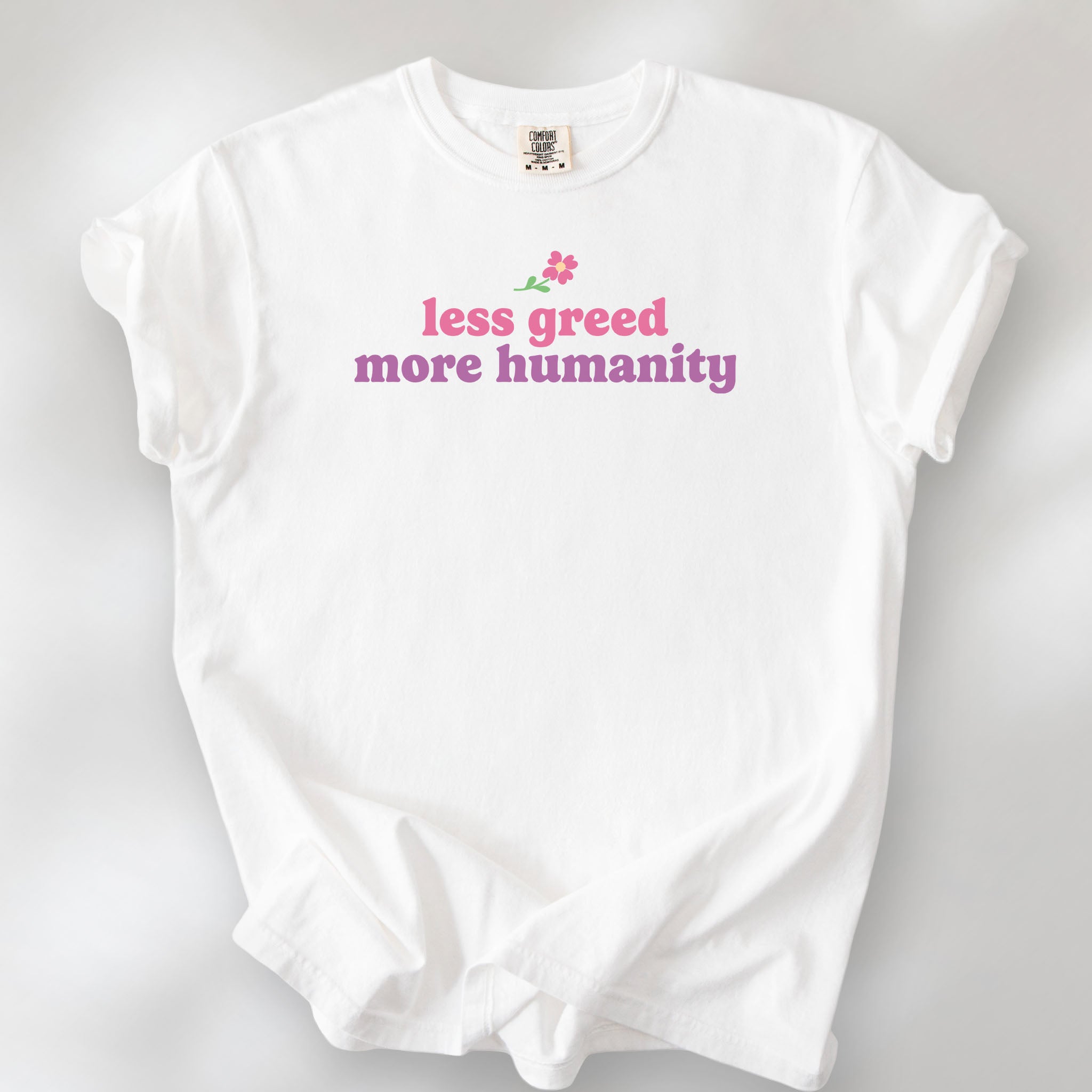 White t-shirt featuring the phrase "Less Greed More Humanity" in soft retro typography with a small flower graphic above the text.