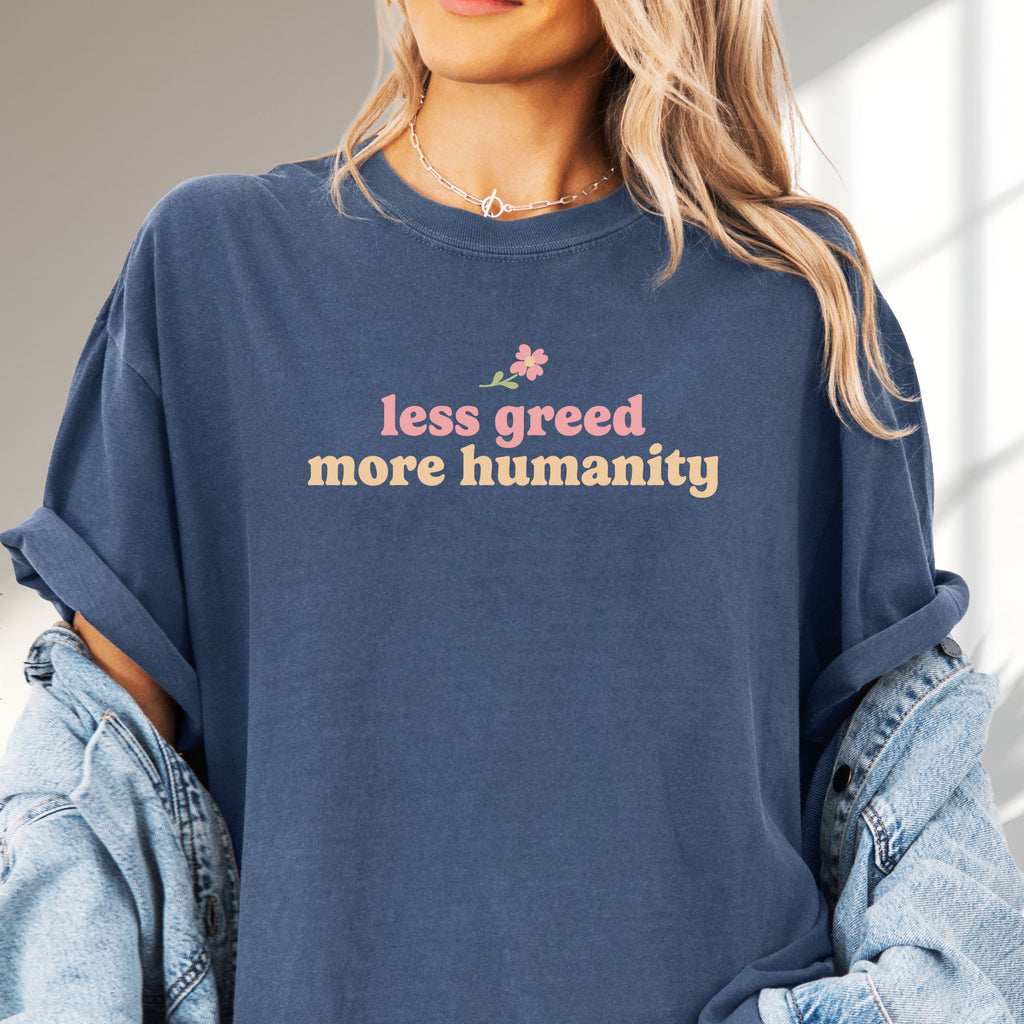 Female model wearing oversized denim t-shirt featuring the phrase "Less Greed More Humanity" in soft retro typography with a small flower graphic above the text.