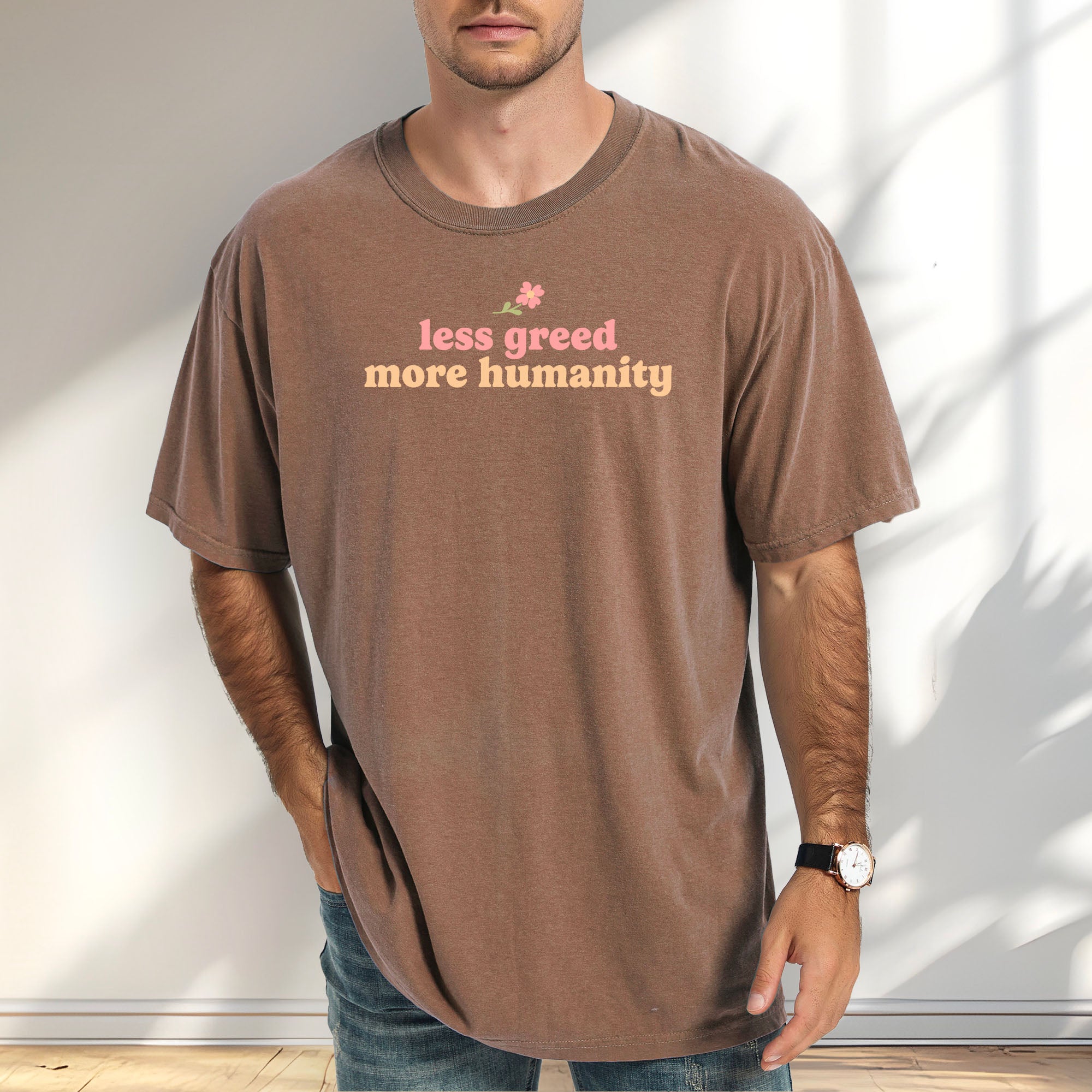 Male model wearing espresso t-shirt featuring the phrase "Less Greed More Humanity" in soft retro typography with a small flower graphic above the text.