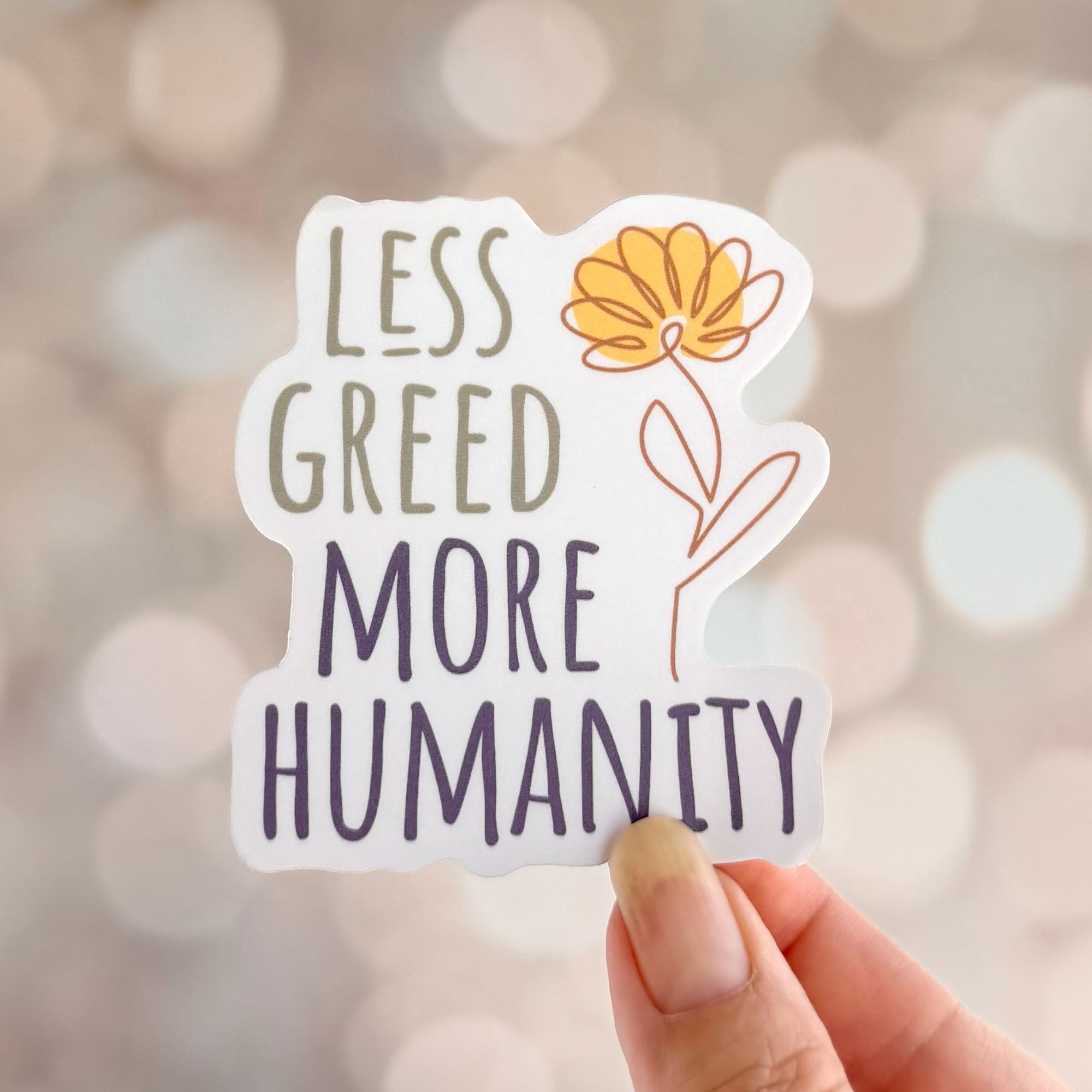 Less Greed More Humanity matte vinyl die-cut sticker with a minimalist yellow flower illustration, shown held in a hand against a soft neutral background.