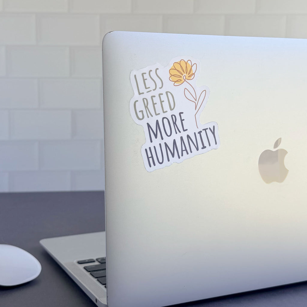 Less Greed More Humanity matte vinyl die-cut sticker with a minimalist yellow flower illustration, shown stuck on an apple laptop computer.
