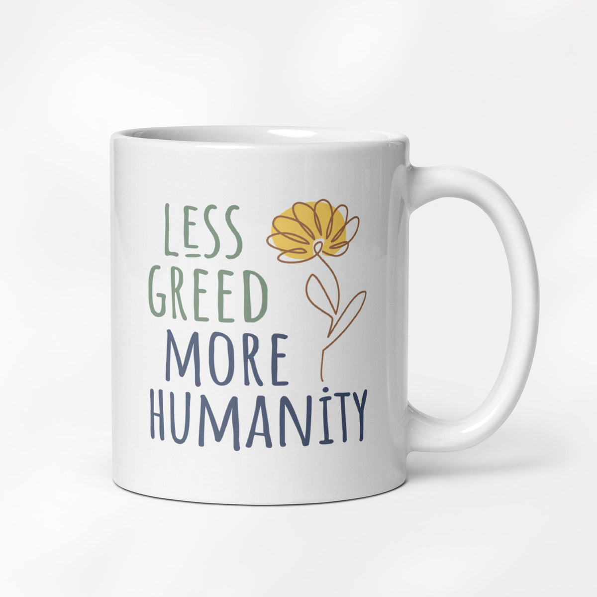 11 ounce white ceramic mug featuring the phrase “Less Greed More Humanity” in hand-lettered typography with green and blue text alongside a simple yellow flower illustration.