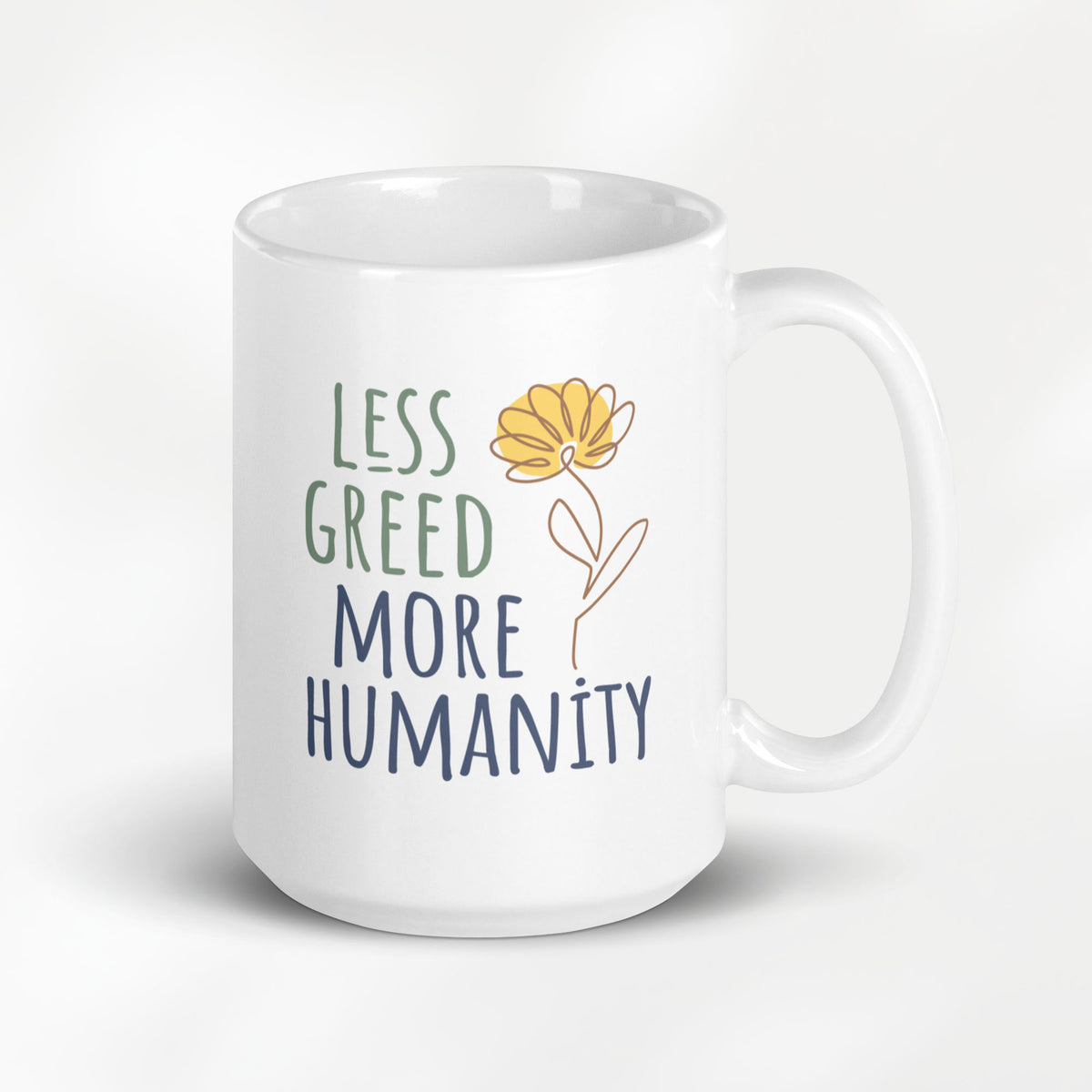 15 ounce white ceramic mug featuring the phrase “Less Greed More Humanity” in hand-lettered typography with green and blue text alongside a simple yellow flower illustration.