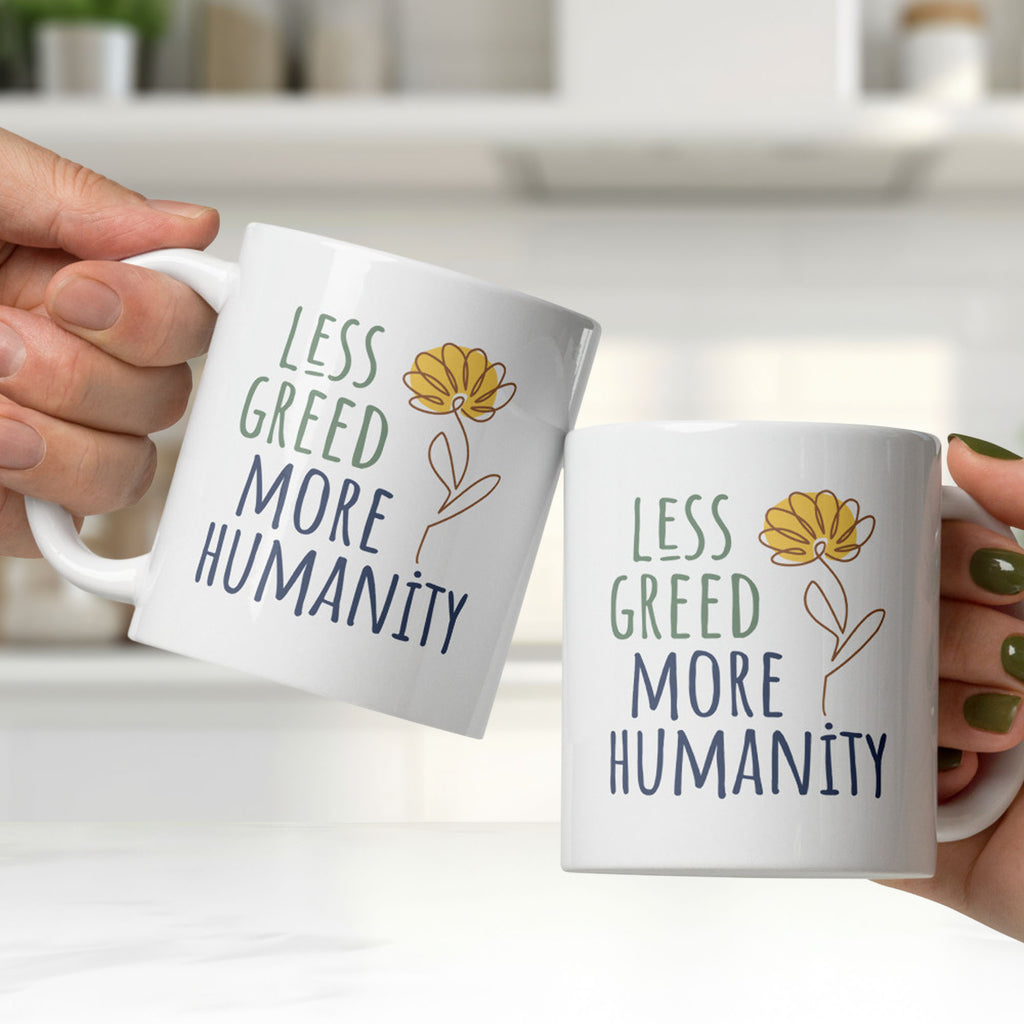Two people holding white ceramic mug featuring the phrase “Less Greed More Humanity” in hand-lettered typography with green and blue text alongside a simple yellow flower illustration, clinking them together in a modern white kitchen.