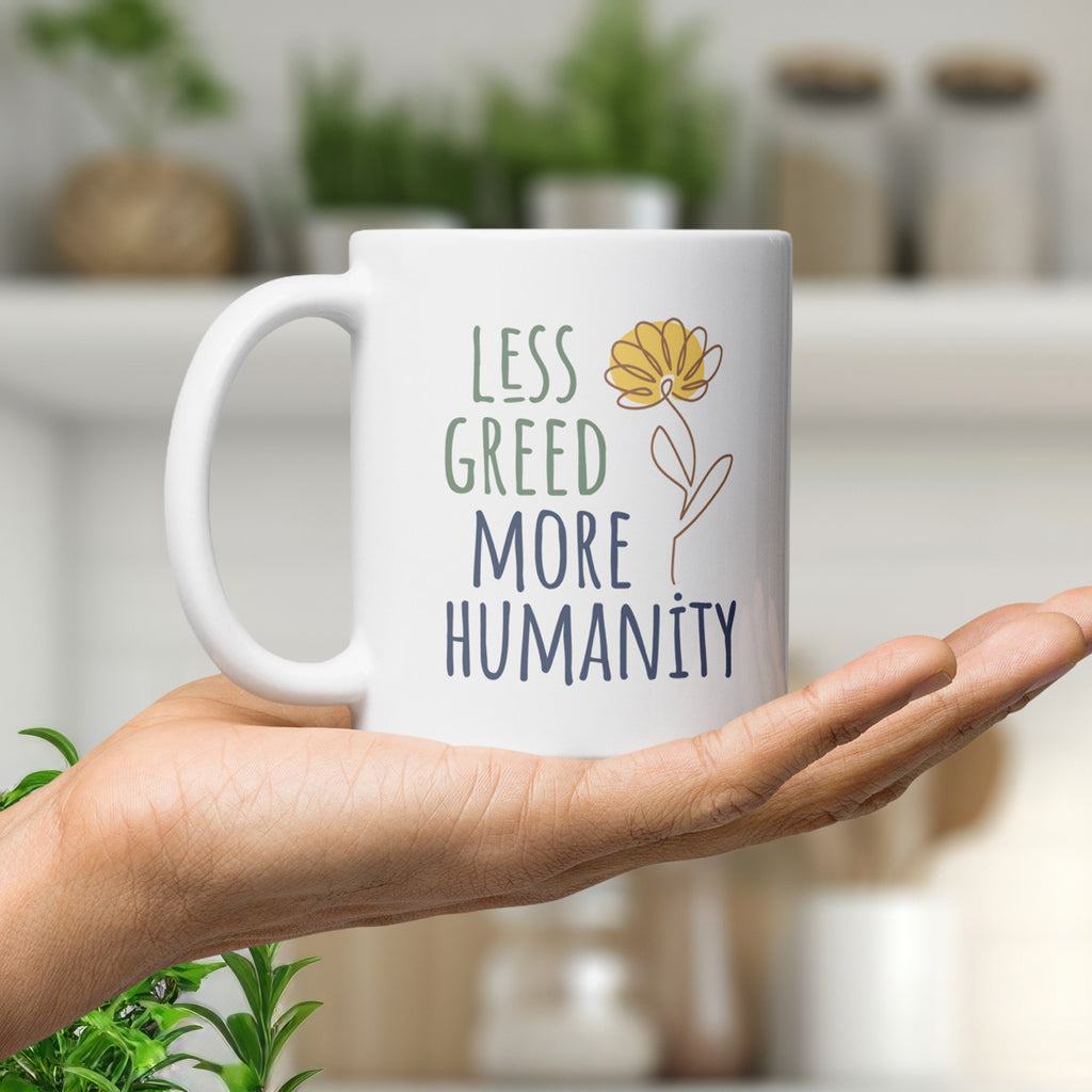 White ceramic mug featuring the phrase “Less Greed More Humanity” in hand-lettered typography with green and blue text alongside a simple yellow flower illustration, held in the palm of a hand in a modern kitchen with plants.