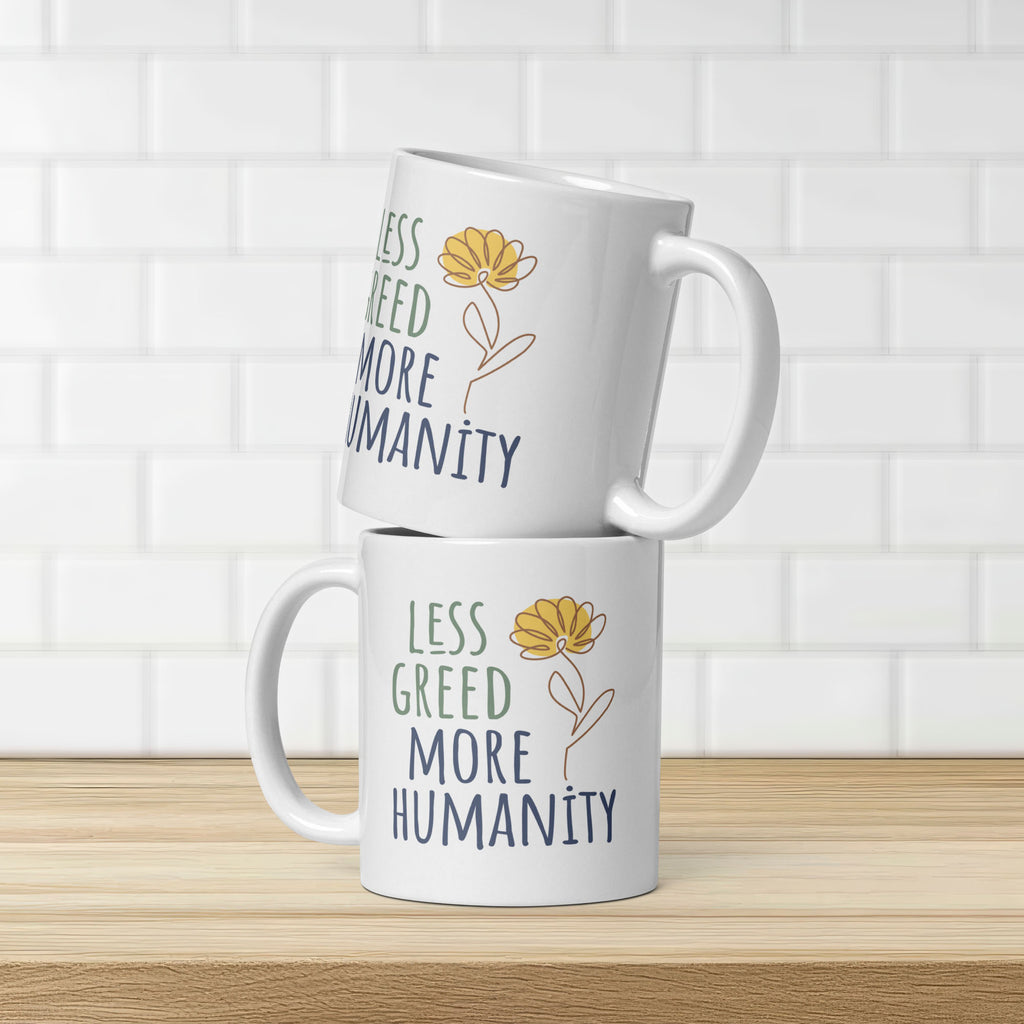 Two stacked white ceramic mug featuring the phrase “Less Greed More Humanity” in hand-lettered typography with green and blue text alongside a simple yellow flower illustration, on a kitchen counter, showing design on both sides of mug.