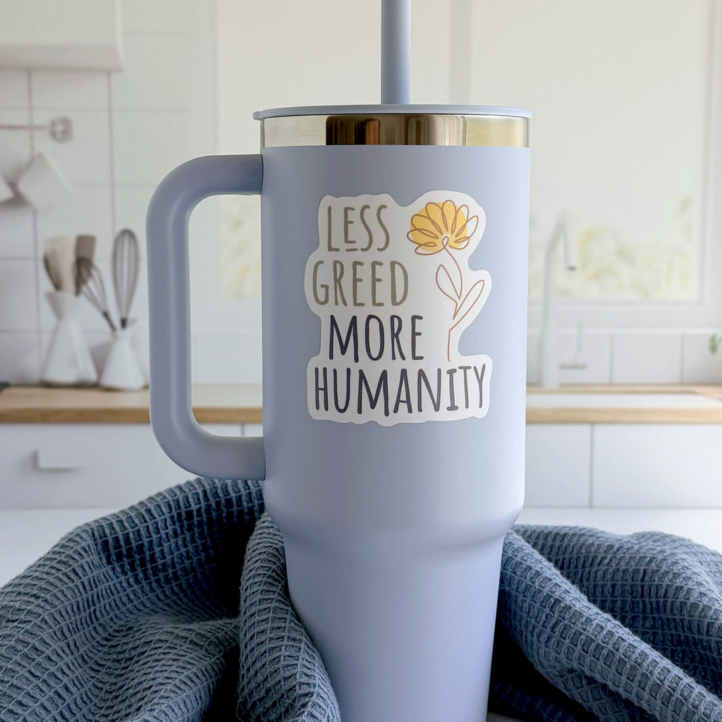 Less Greed More Humanity matte vinyl die-cut sticker with a minimalist yellow flower illustration, on a tumbler in a kitchen.
