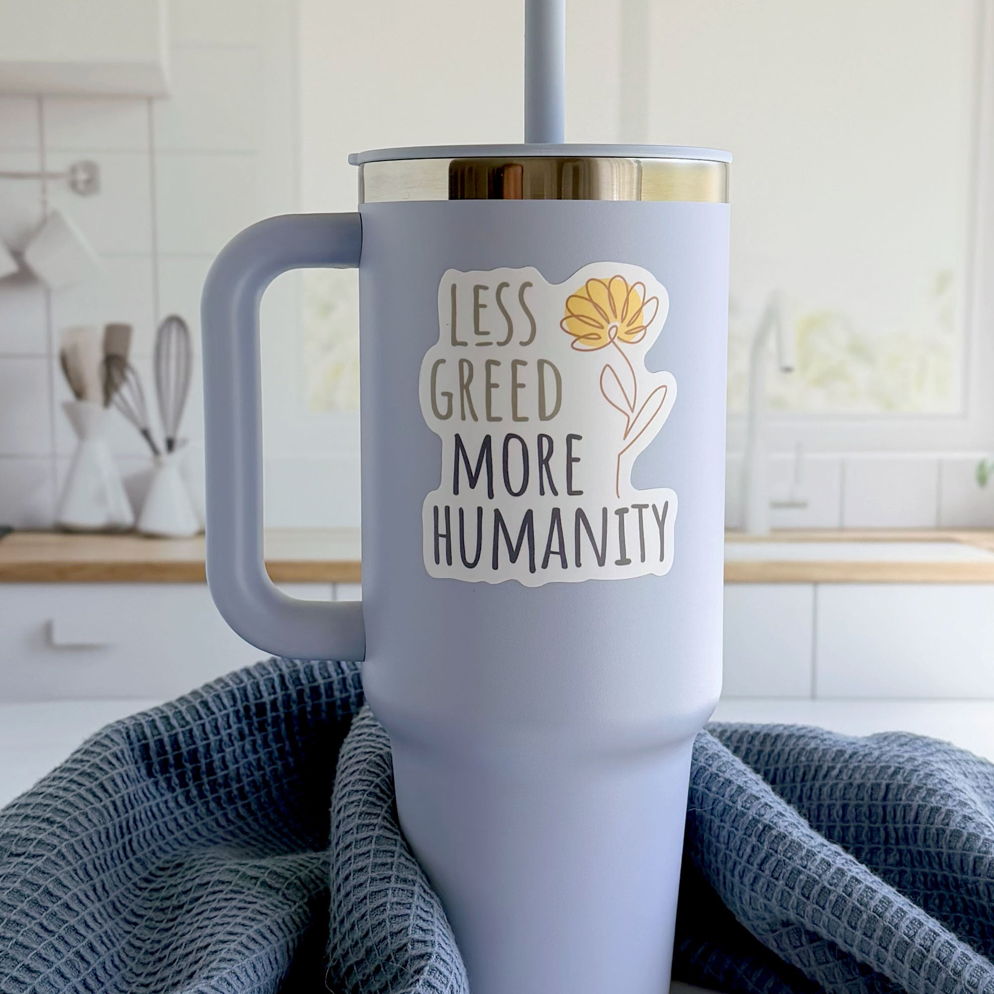 Less Greed More Humanity matte vinyl die-cut sticker with a minimalist yellow flower illustration, on a tumbler in a kitchen.