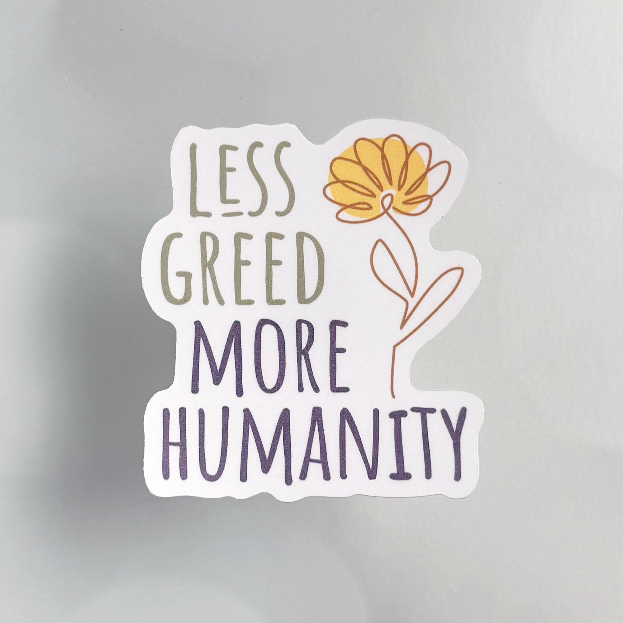 Less Greed More Humanity matte vinyl die-cut sticker with a minimalist yellow flower illustration.