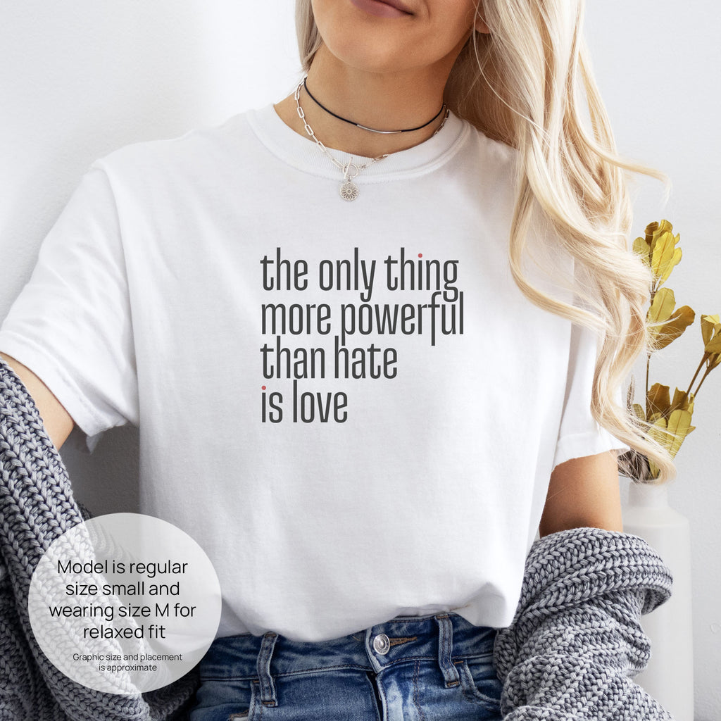 Female model wearing white t-shirt with a stylized text block reading “the only thing more powerful than hate is love." Includes size and fit information for the model who is regular size small, and wearing a size M shirt for a relaxed fit.
