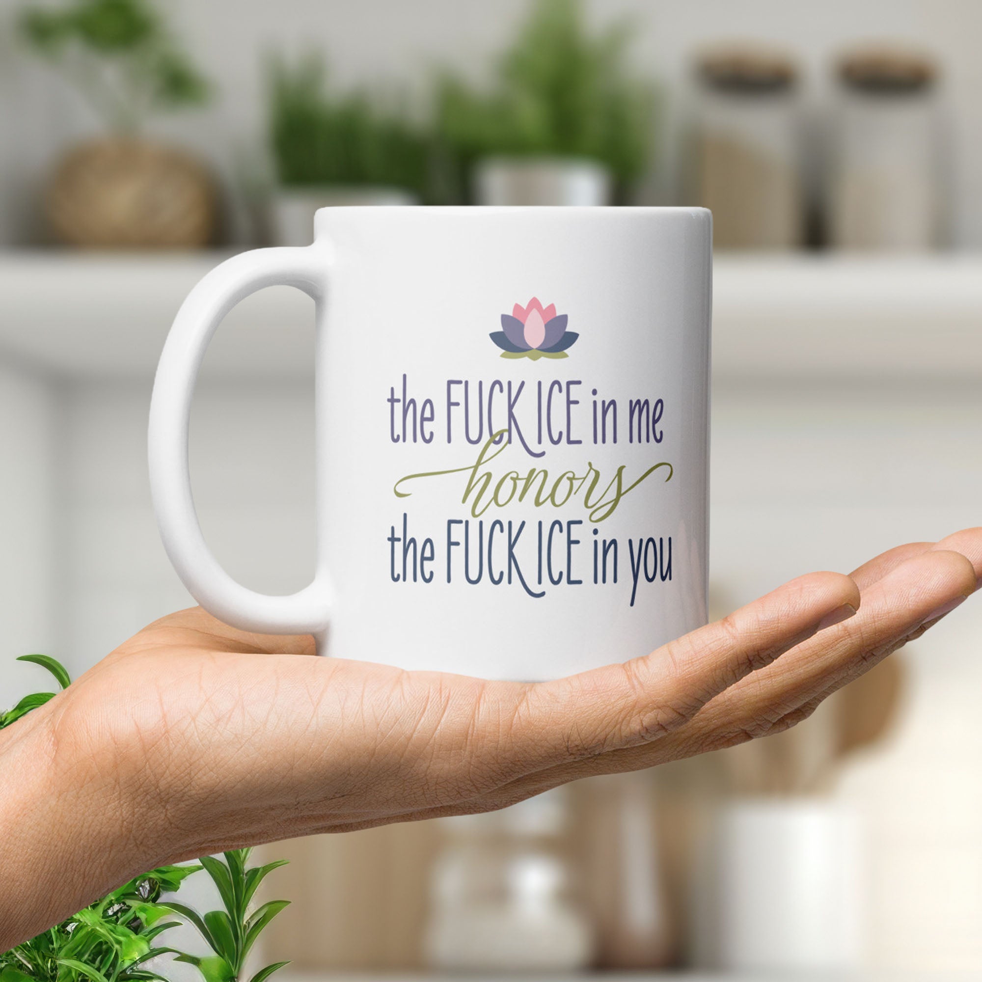 White ceramic coffee mug featuring the phrase "The Fck ICE in Me Honors the Fck ICE in You" in decorative typography with a lotus flower above, resting on the palm of a hand in a modern kitchen.
