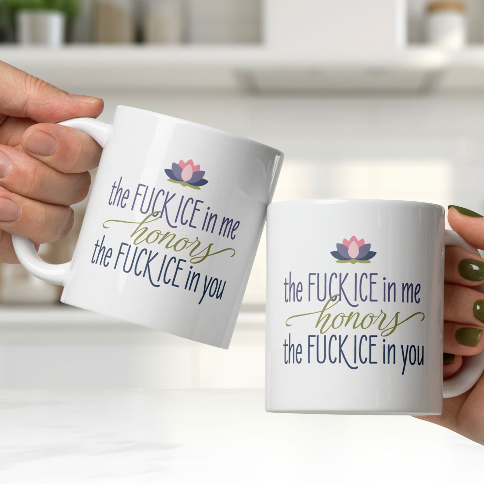 Two white ceramic coffee mugs being clicked together, featuring the phrase "The Fck ICE in Me Honors the Fck ICE in You" in decorative typography with a lotus flower above, in a clean white kitchen.