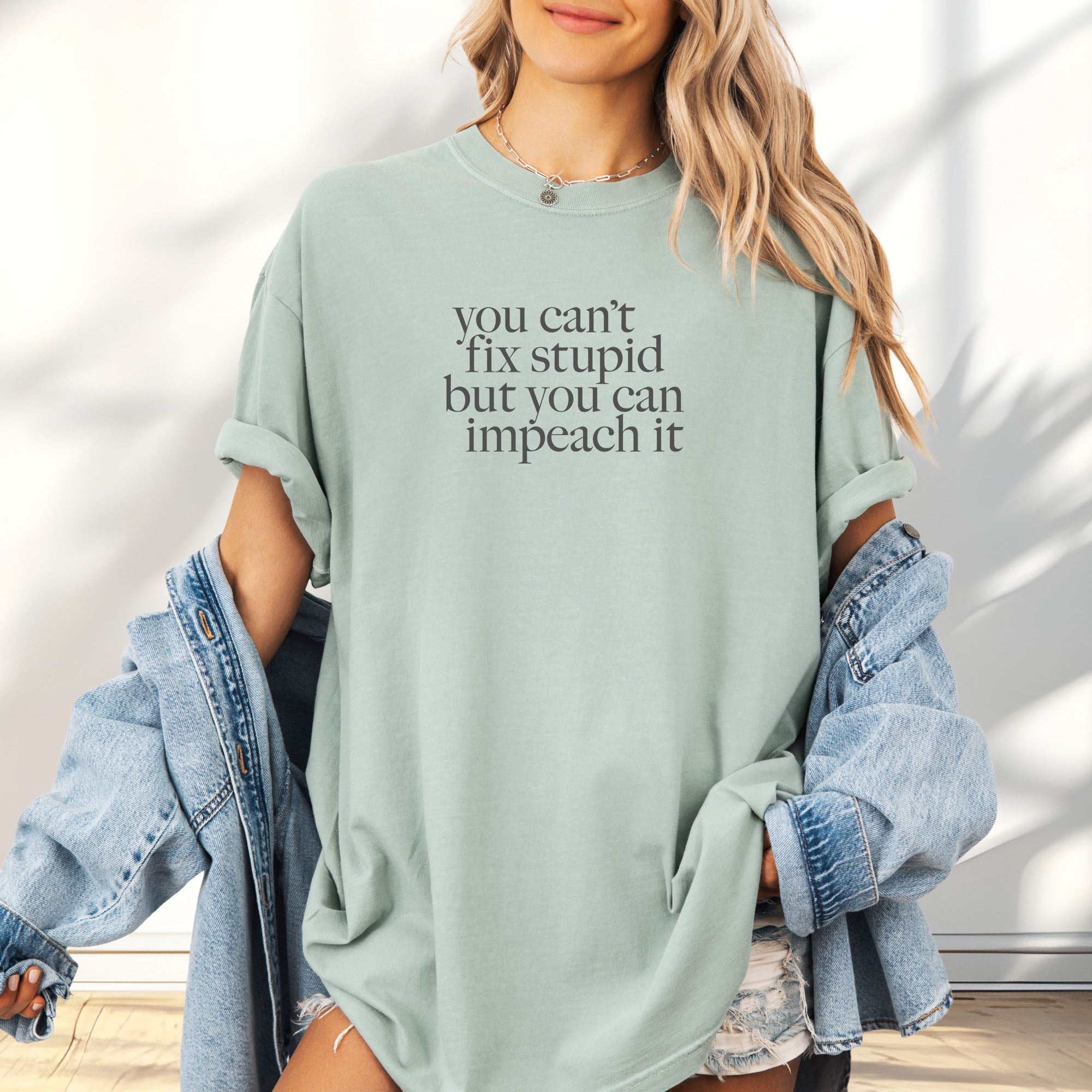 Female model wearing bay political t-shirt with bold gray text reading “You can’t fix stupid but you can impeach it,” minimalist anti-Trump protest design.