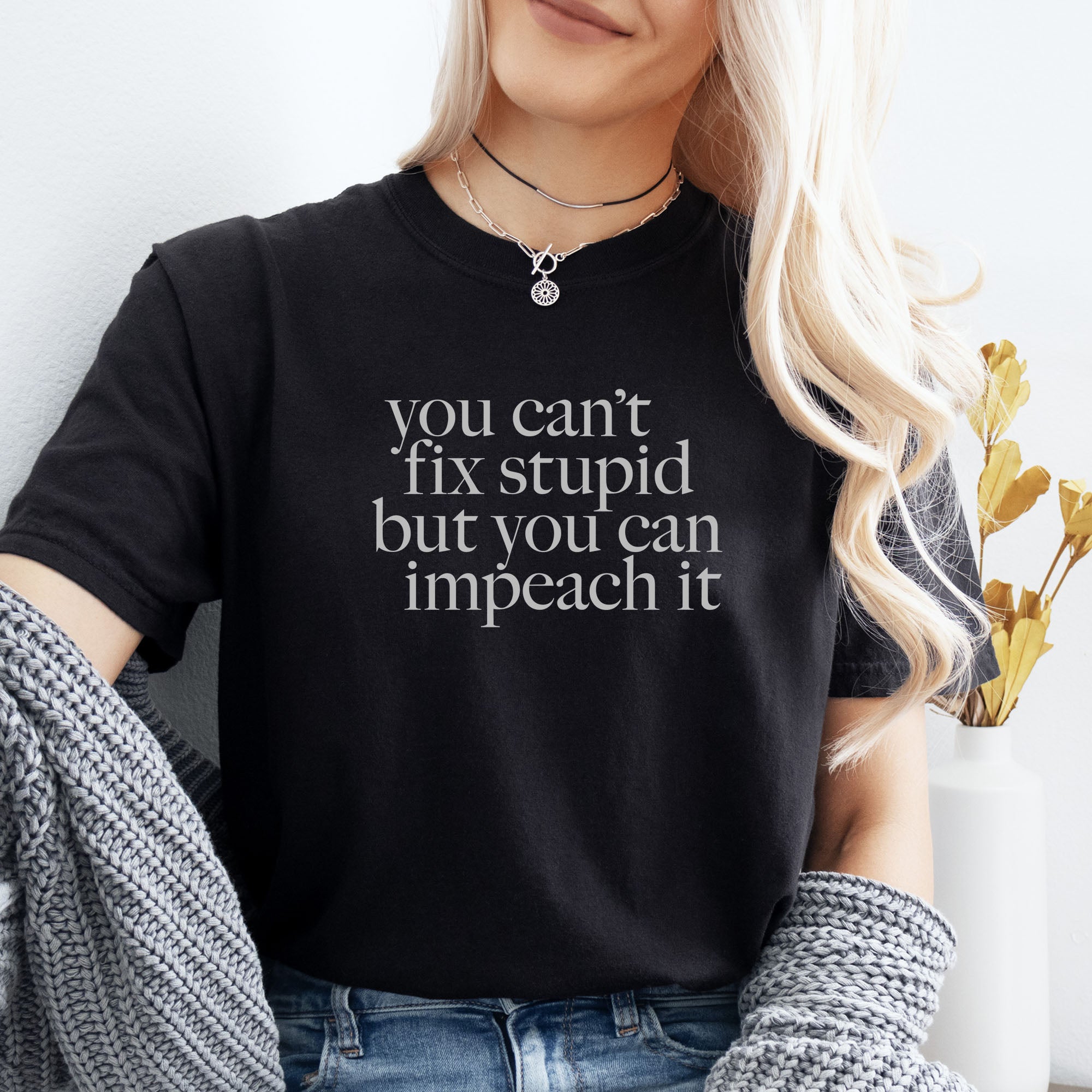 Female model wearing black political t-shirt with light grey serif text reading “You can’t fix stupid but you can impeach it,” minimalist anti-Trump protest design.