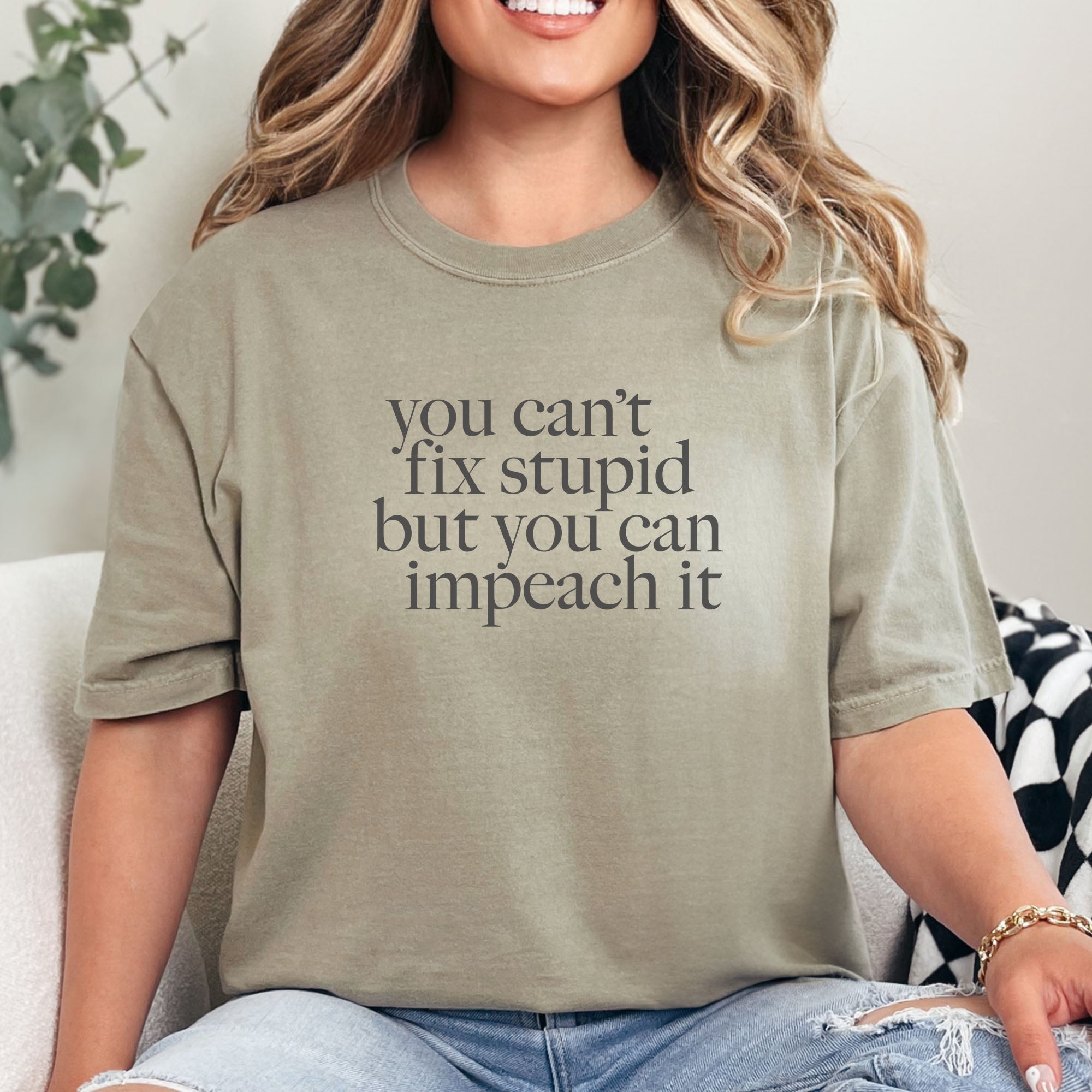 Female model wearing khaki political t-shirt with bold gray text reading “You can’t fix stupid but you can impeach it,” minimalist anti-Trump protest design.