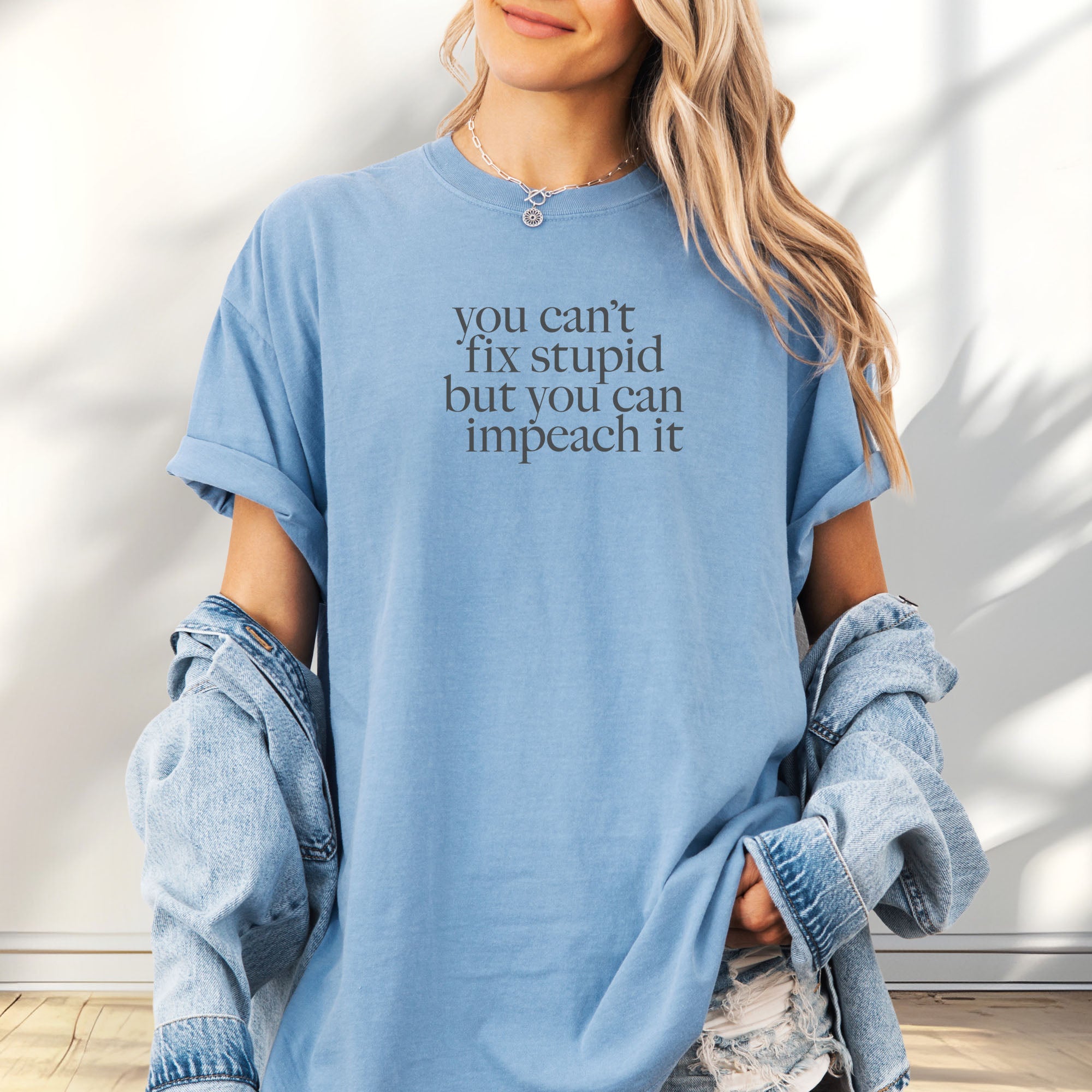 Female model wearing oversized washed denim political t-shirt with bold gray text reading “You can’t fix stupid but you can impeach it,” minimalist anti-Trump protest design.