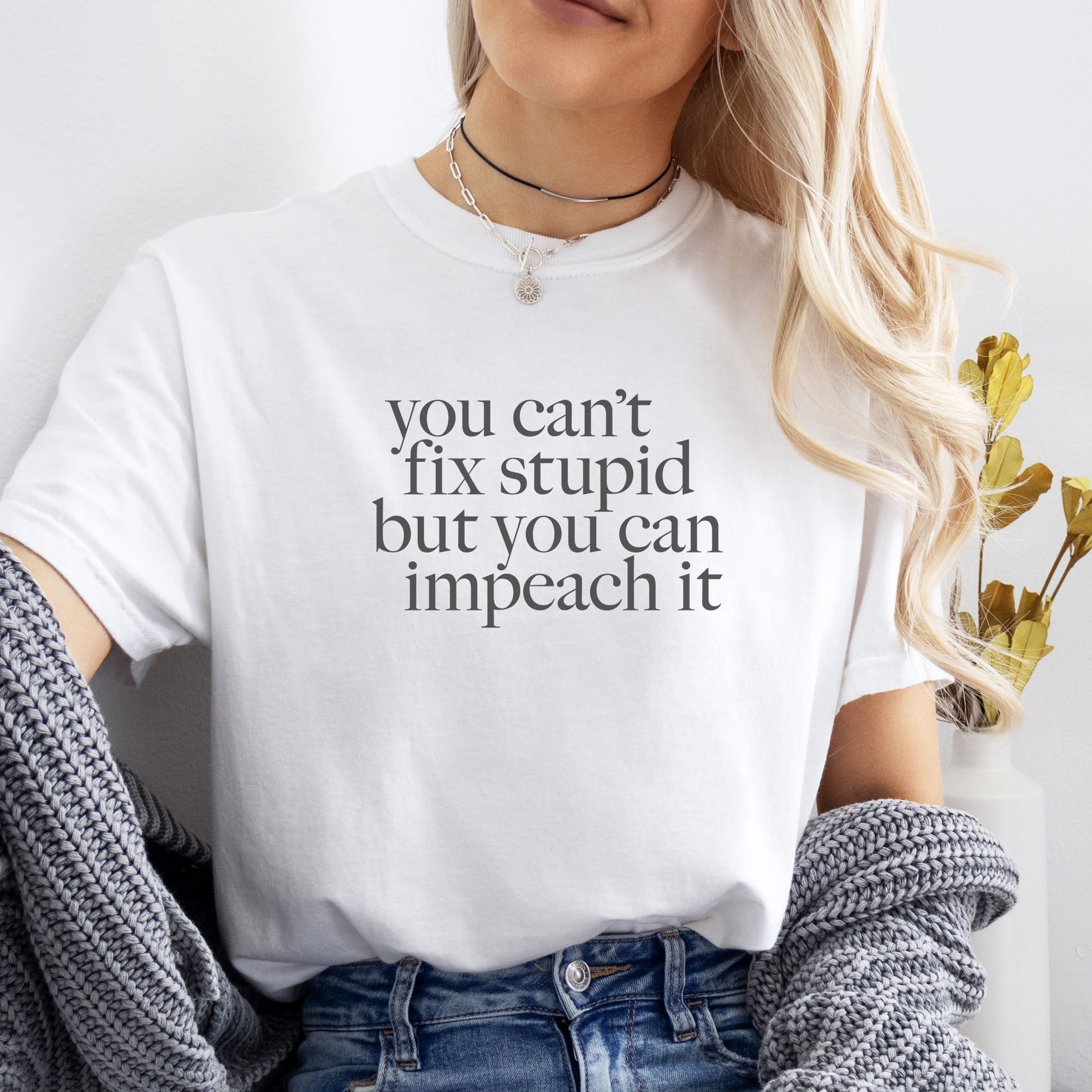 Female model wearing white political t-shirt with bold gray text reading “You can’t fix stupid but you can impeach it,” minimalist anti-Trump protest design.