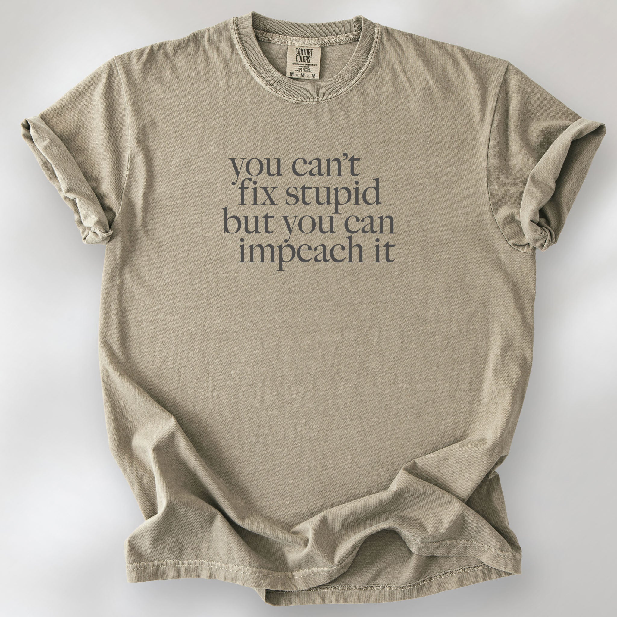 Khaki political t-shirt with bold gray text reading “You can’t fix stupid but you can impeach it,” minimalist anti-Trump protest design.