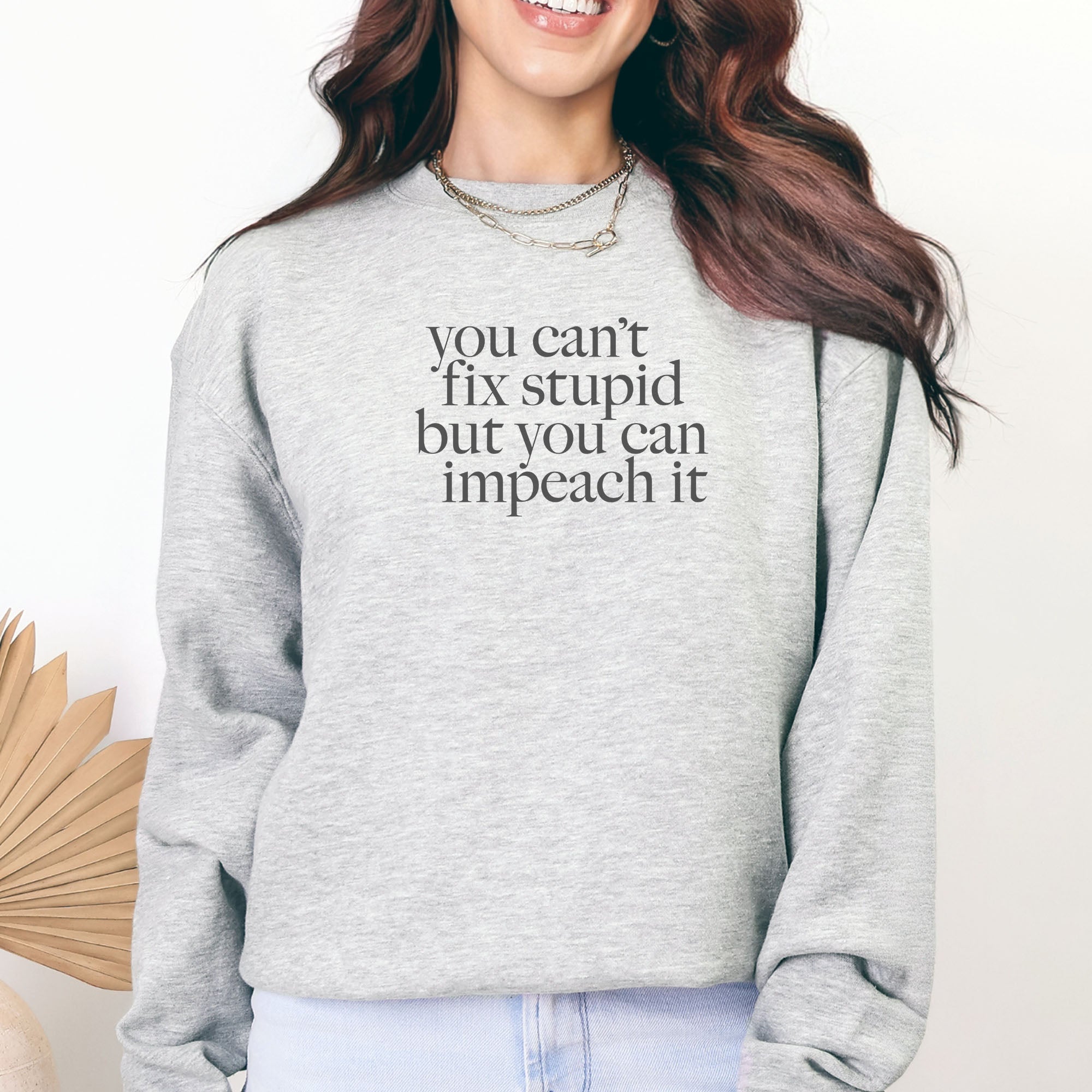 Female model wearing grey political sweatshirt shirt with styled serif text block reading “You can’t fix stupid but you can impeach it,” minimalist anti-Trump protest design.