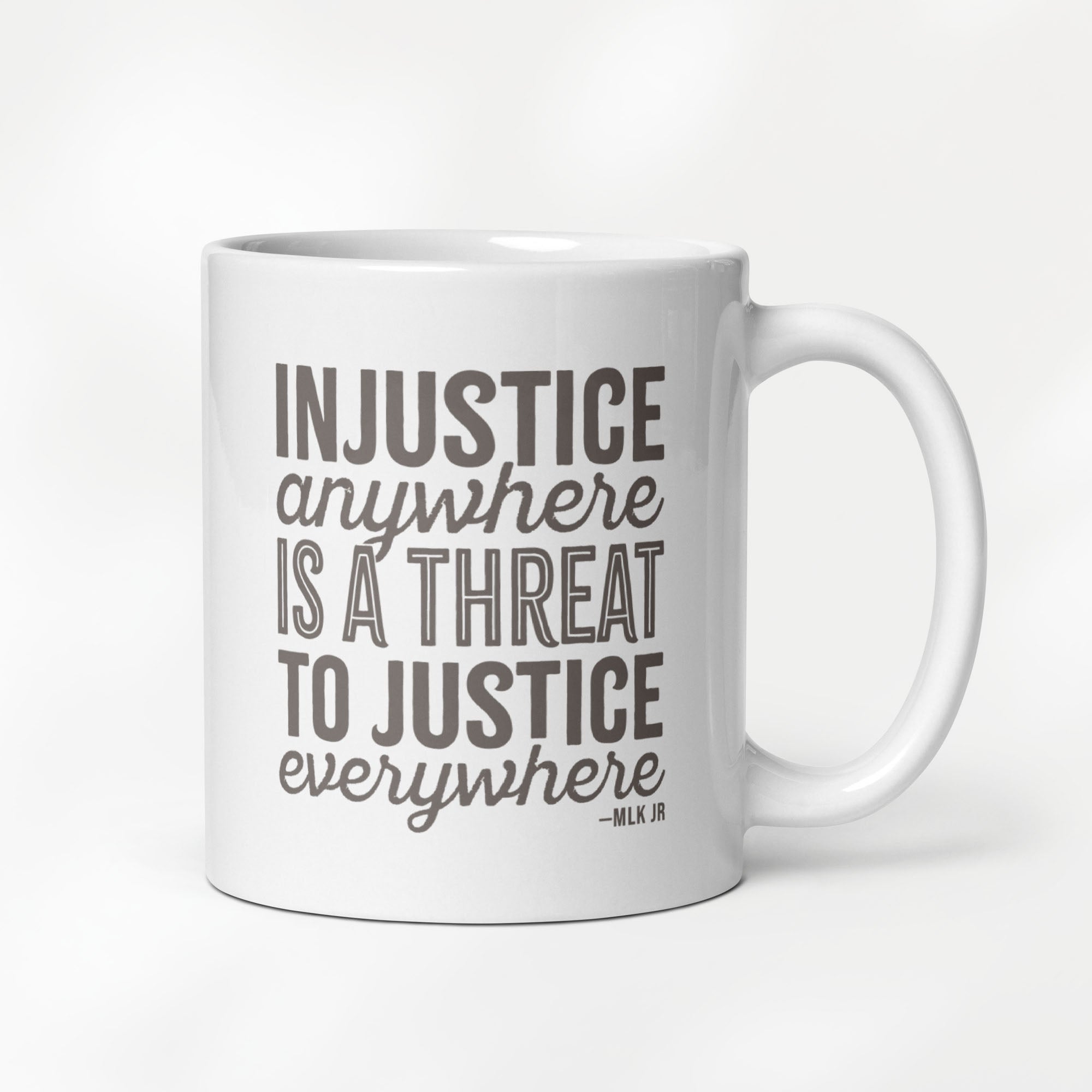 11 ounce white ceramic mug with bold text graphic reading “injustice anywhere is a threat to justice everywhere,” a quote from MLK Junior from Letters from Birmingham Jail, on a desk with a plant and a keyboard.
