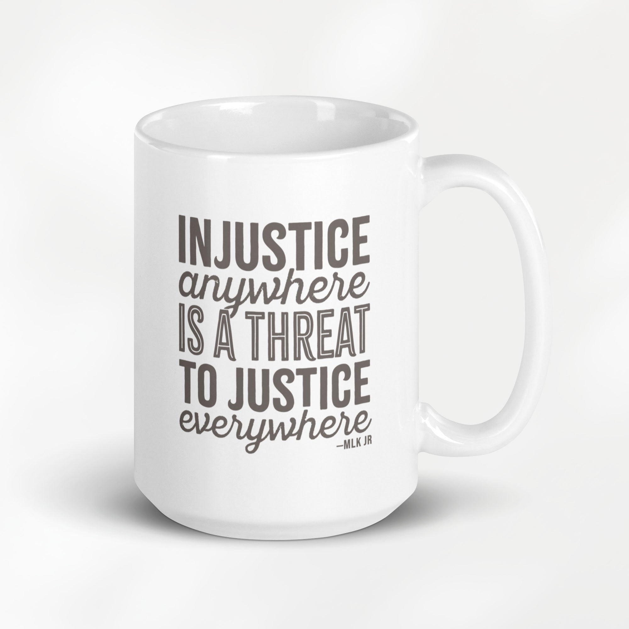 15 ounce white ceramic mug with bold text graphic reading “injustice anywhere is a threat to justice everywhere,” a quote from MLK Junior from Letters from Birmingham Jail, on a desk with a plant and a keyboard.