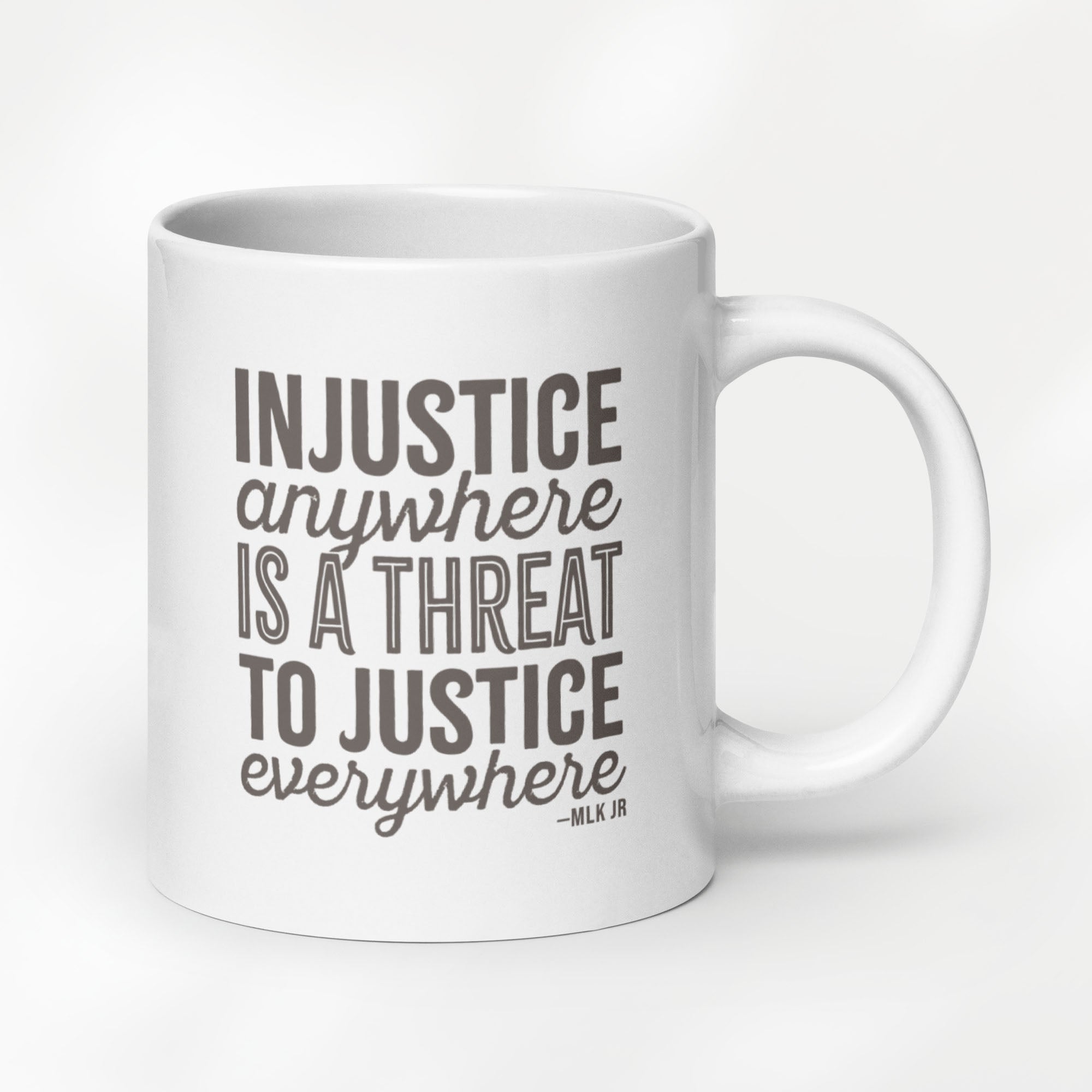 20 ounce white ceramic mug with bold text graphic reading “injustice anywhere is a threat to justice everywhere,” a quote from MLK Junior from Letters from Birmingham Jail, on a desk with a plant and a keyboard.