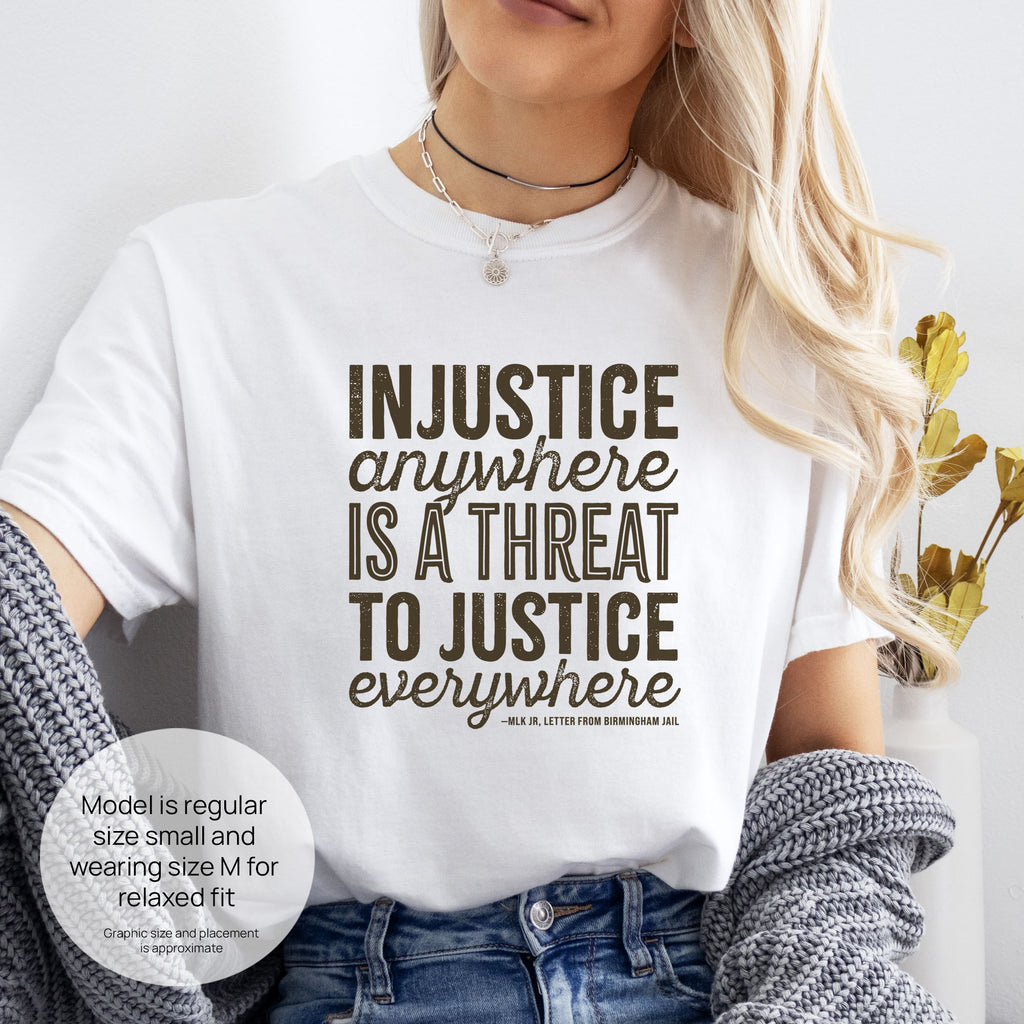 Female model wearing white t-shirt with bold text reading “Injustice anywhere is a threat to justice everywhere” quote from Martin Luther King Jr, MLK Jr, Letter from Birmingham jail — civil rights protest apparel for activists. Includes size and fit information for the model who is regular size small, and wearing a size M shirt for a relaxed fit.
