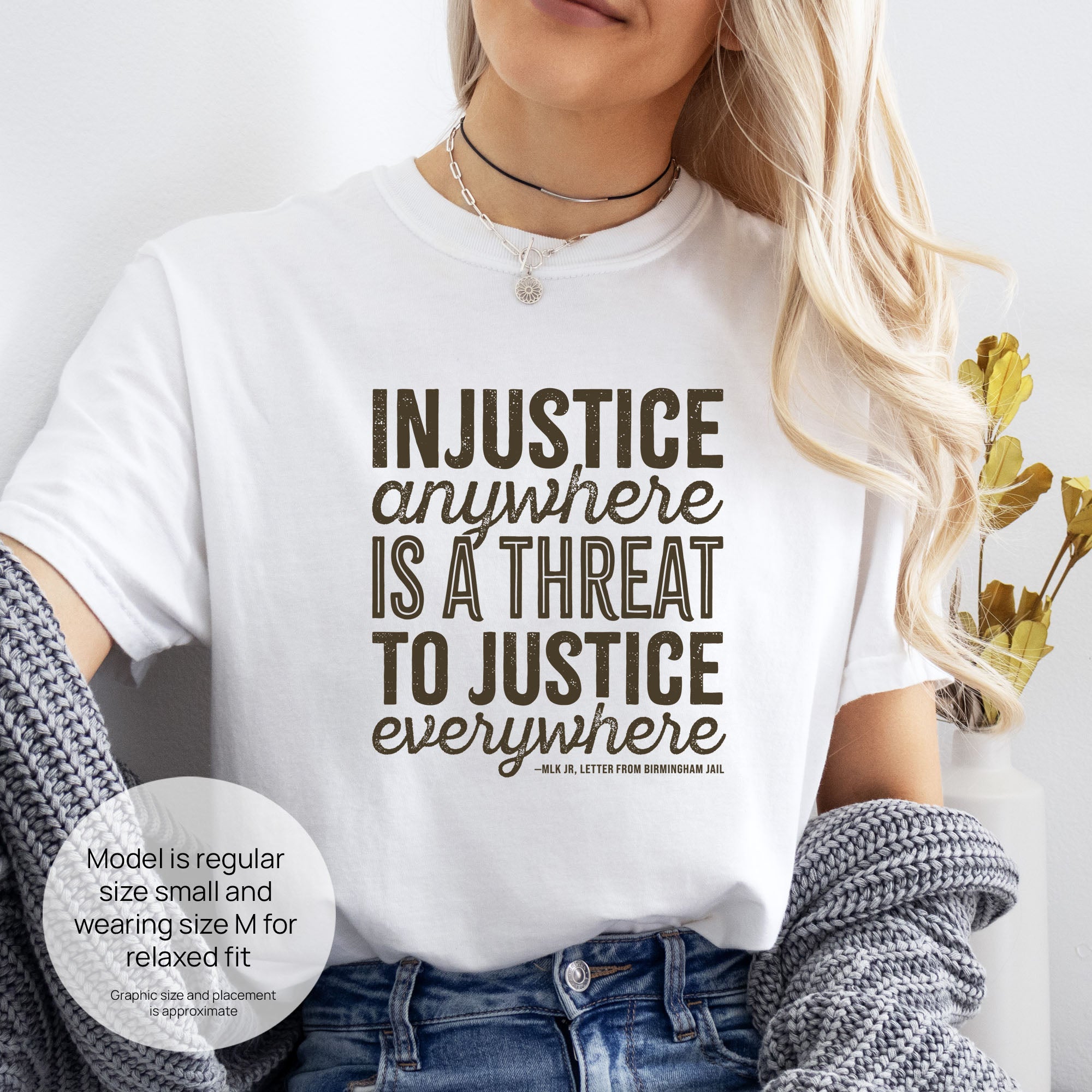 Female model wearing white t-shirt with bold text reading “Injustice anywhere is a threat to justice everywhere” quote from Martin Luther King Jr, MLK Jr, Letter from Birmingham jail — civil rights protest apparel for activists. Includes size and fit information for the model who is regular size small, and wearing a size M shirt for a relaxed fit.
