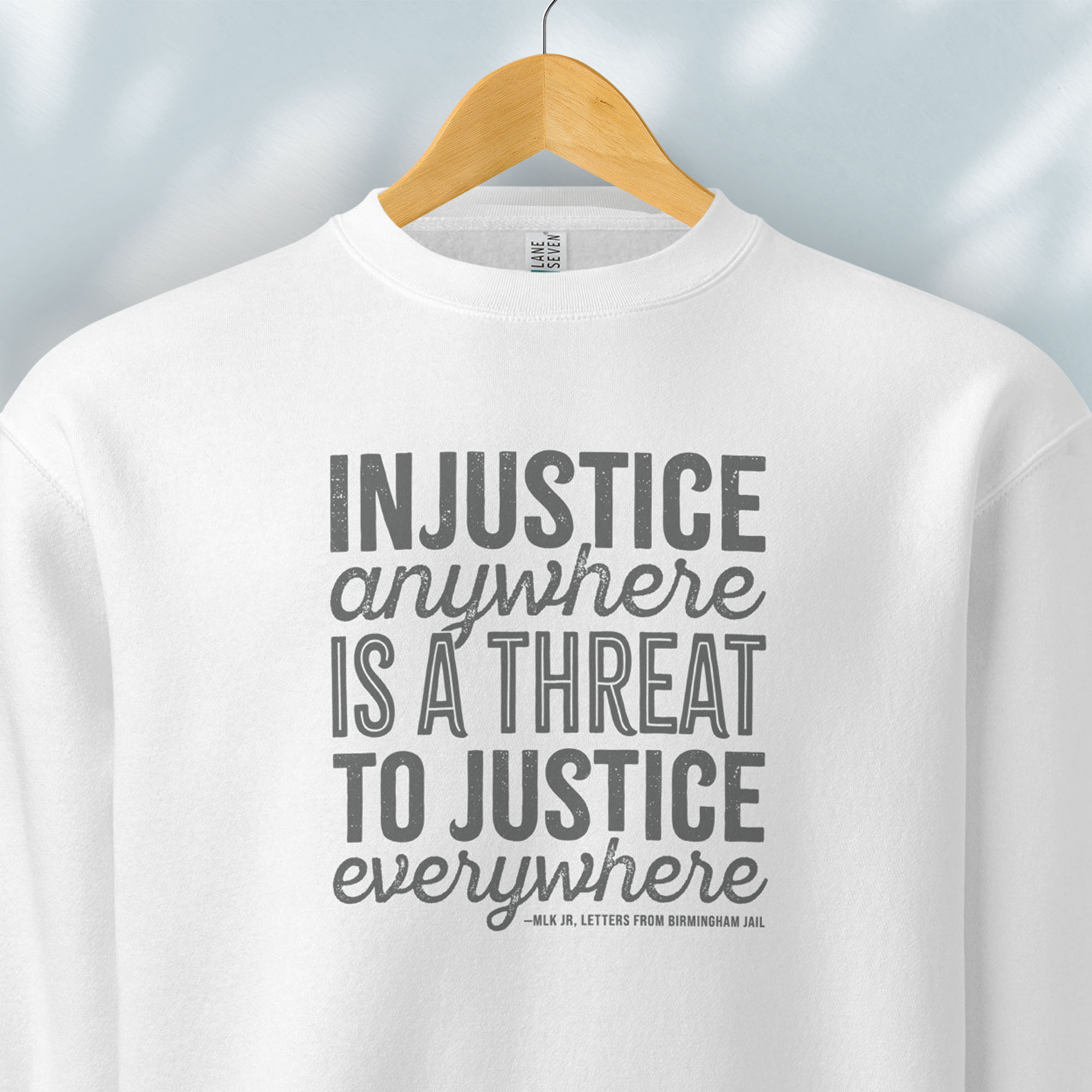 Hanging white sweatshirt with bold text reading “Injustice anywhere is a threat to justice everywhere” quote from Martin Luther King Jr, MLK Jr, Letters from Birmingham jail — civil rights protest apparel for activists.
