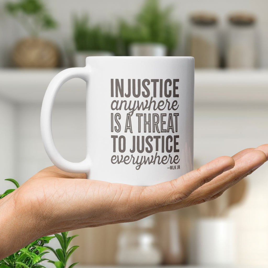 White ceramic mug with bold text graphic reading “injustice anywhere is a threat to justice everywhere,” a quote from MLK Junior from Letters from Birmingham Jail, in the palm of a person's hand.
