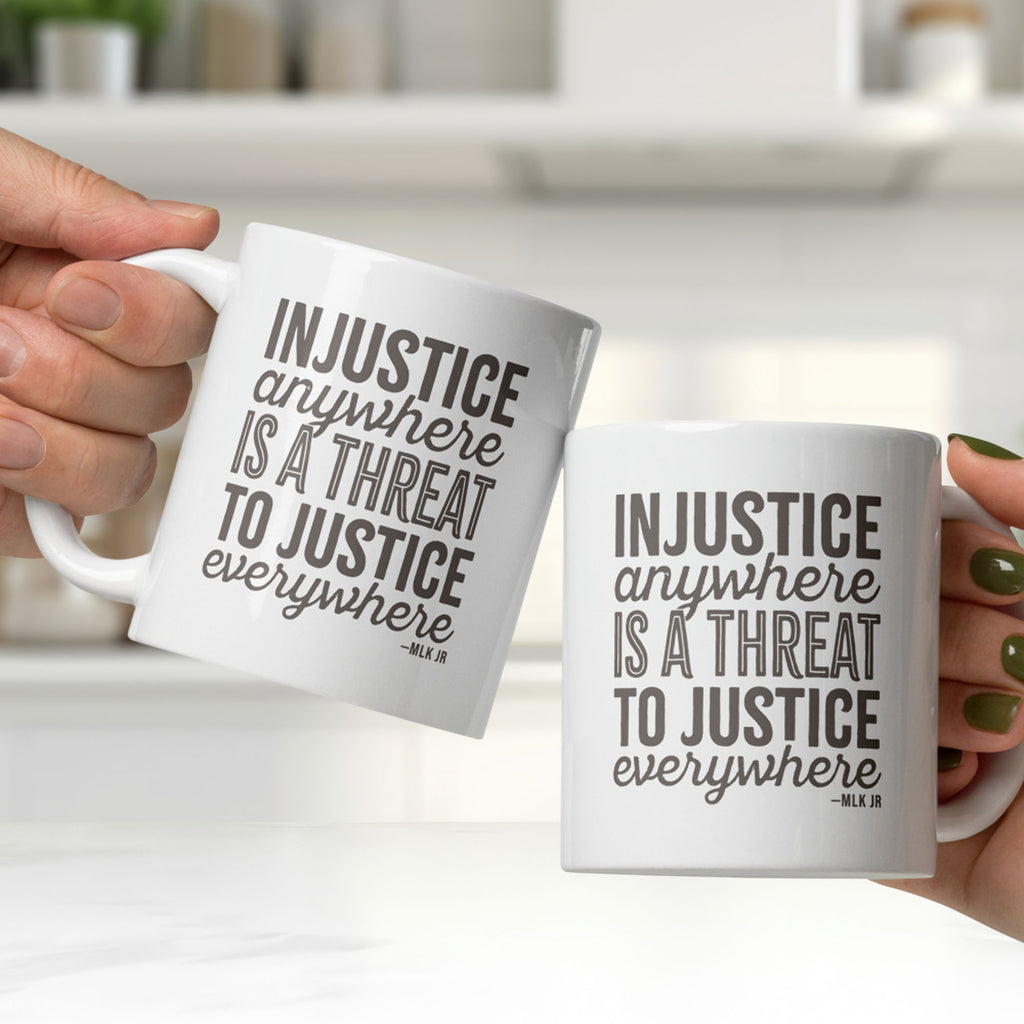 Two hands, each holding a ceramic mug with bold text graphic reading “injustice anywhere is a threat to justice everywhere,” a quote from MLK Junior from Letters from Birmingham Jail, clinking them together in a bright white kitchen.