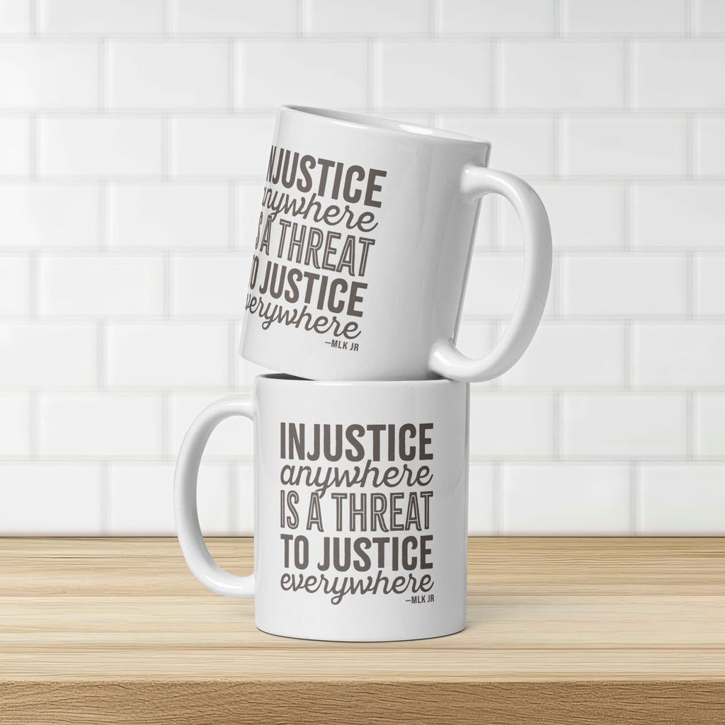 Two white ceramic mugs with bold text graphic reading “injustice anywhere is a threat to justice everywhere,” a quote from MLK Junior from Letters from Birmingham Jail, stacked on a kitchen counter showing both sides of the mug.
