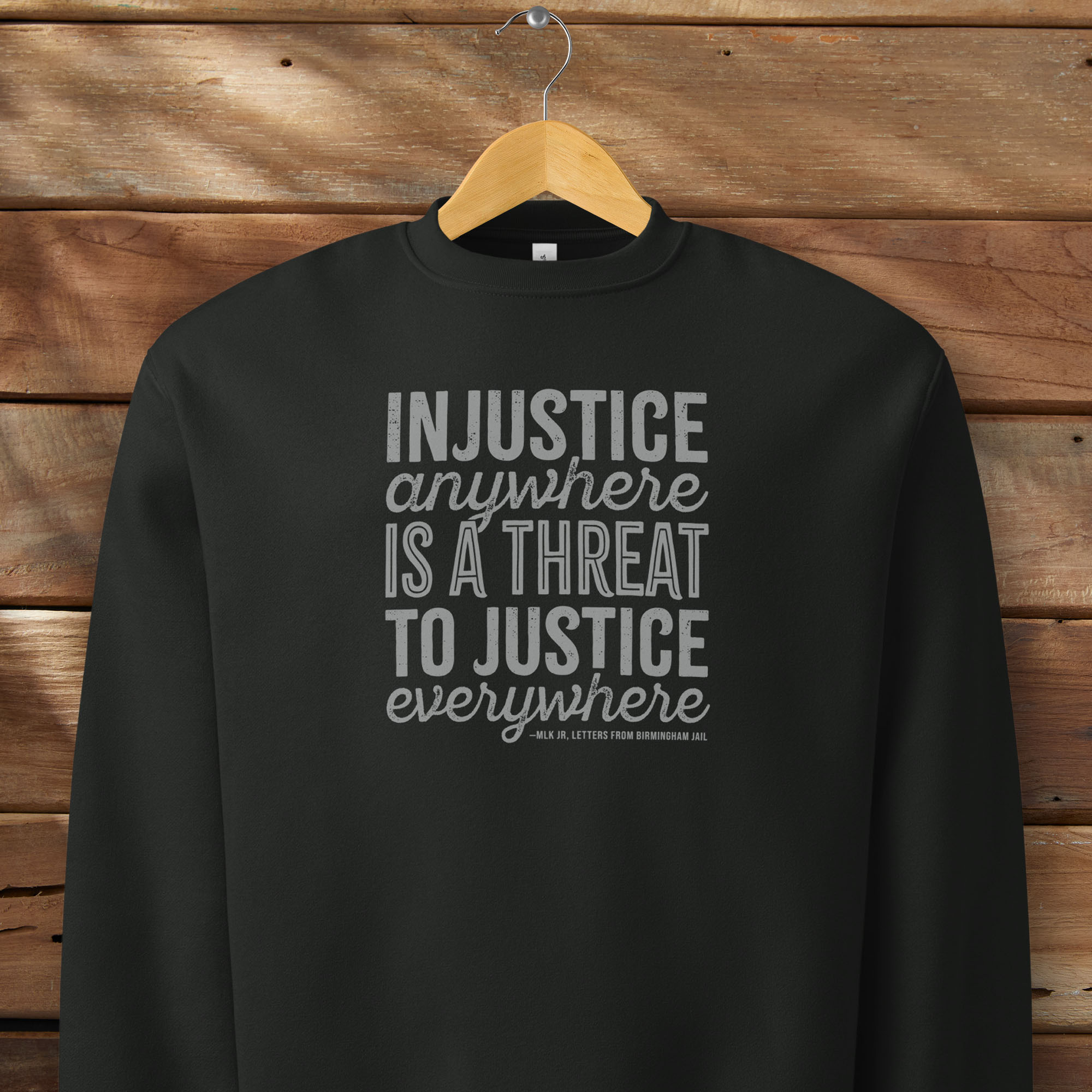 Black sweatshirt with bold text reading “Injustice anywhere is a threat to justice everywhere” quote from Martin Luther King Jr, MLK Jr, Letters from Birmingham jail — civil rights protest apparel for activists. Hanging on a hanger.