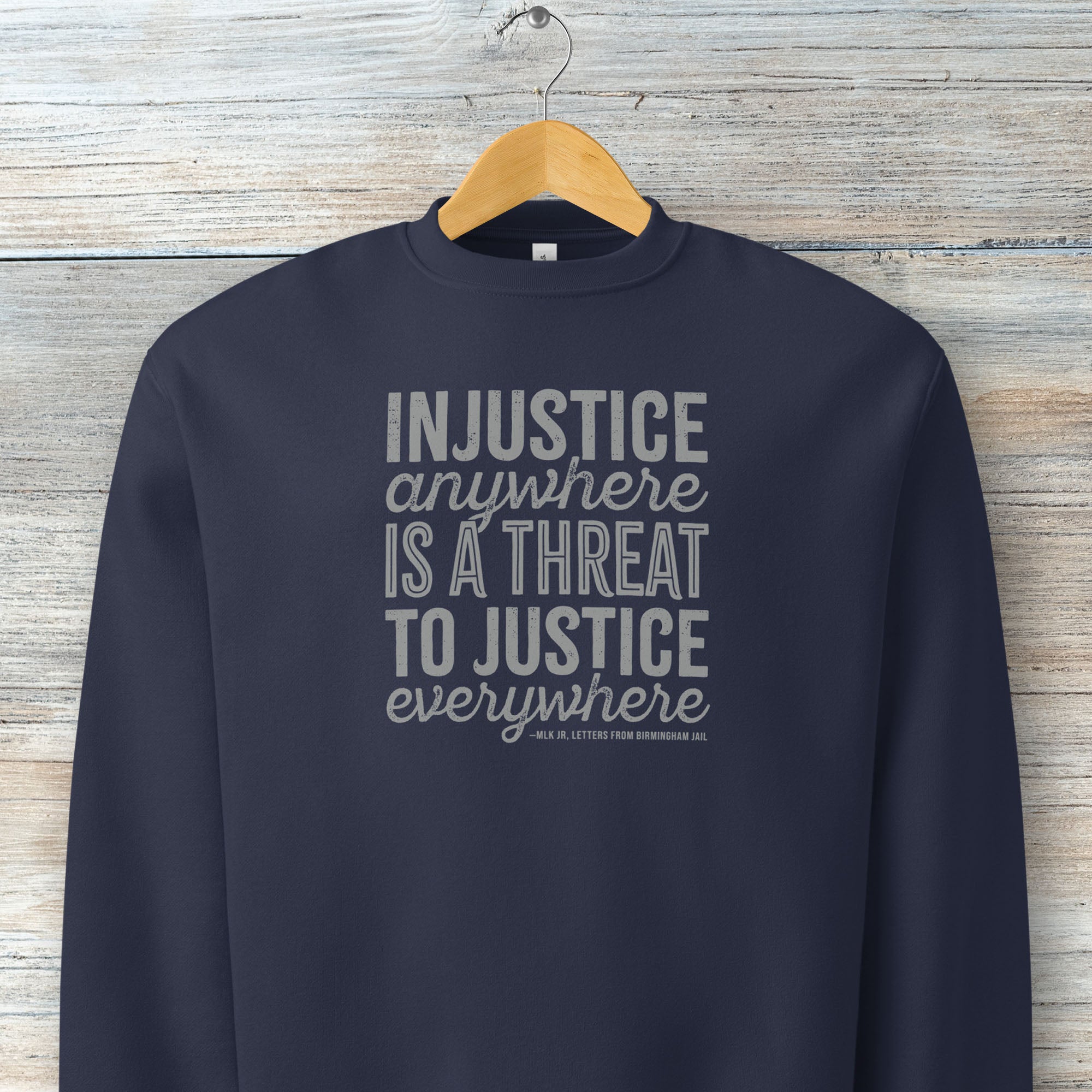 Navy sweatshirt with bold text reading “Injustice anywhere is a threat to justice everywhere” quote from Martin Luther King Jr, MLK Jr, Letters from Birmingham jail — civil rights protest apparel for activists. Hanging on a hanger.