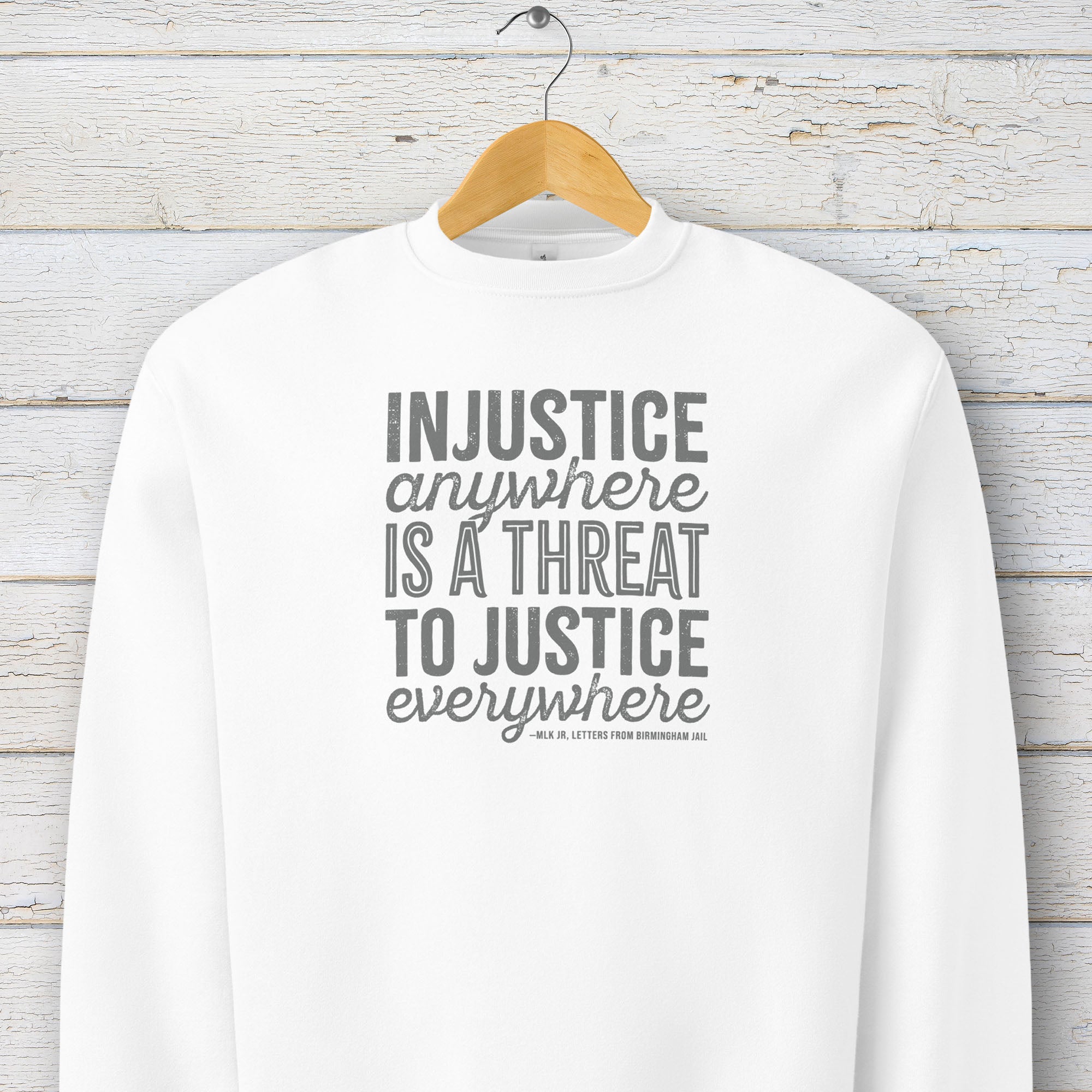 White sweatshirt with bold text reading “Injustice anywhere is a threat to justice everywhere” quote from Martin Luther King Jr, MLK Jr, Letters from Birmingham jail — civil rights protest apparel for activists. Hanging on a hanger.