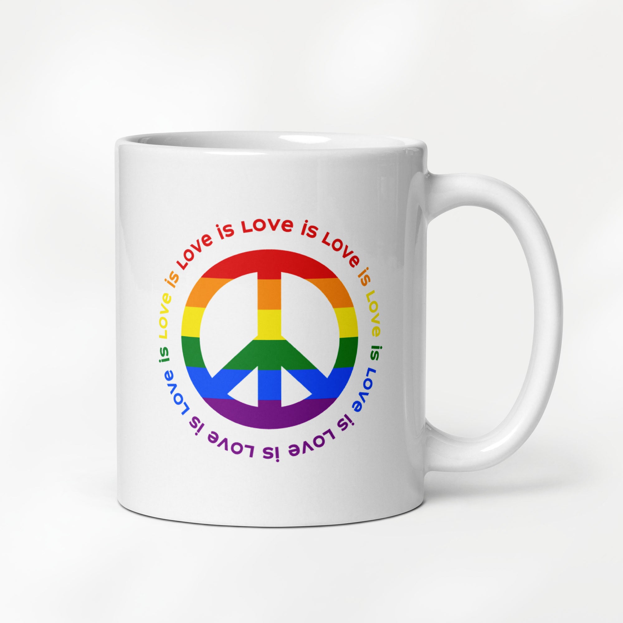 11 ounce white ceramic coffee mug with a peace sign in the colors of the LGBTQ+ flag, surrounded by repeated "love is love" text. On a desk with a keyboard, plant, and book.
