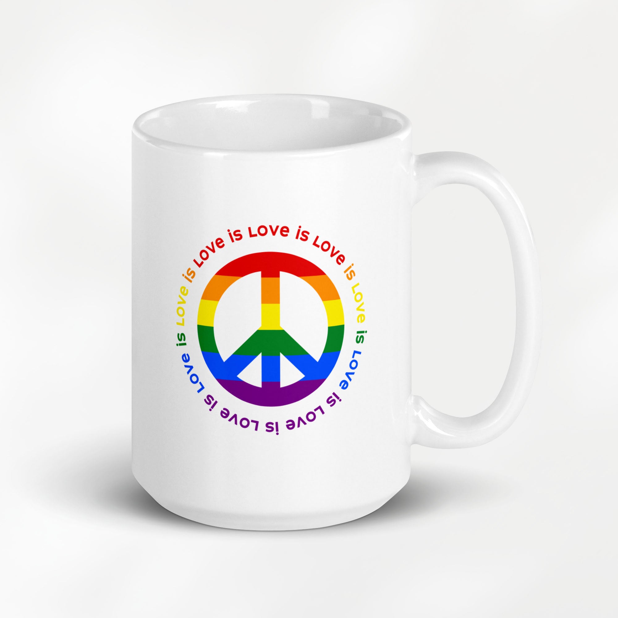 15 ounce white ceramic coffee mug with a peace sign in the colors of the LGBTQ+ flag, surrounded by repeated "love is love" text. On a desk with a keyboard, plant, and book.