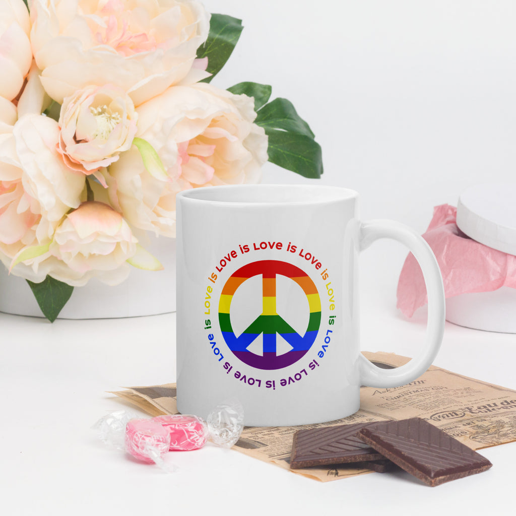 White ceramic coffee mug with a peace sign in the colors of the LGBTQ+ flag, surrounded by repeated "love is love" text. On a tabletop with flowers and candy.