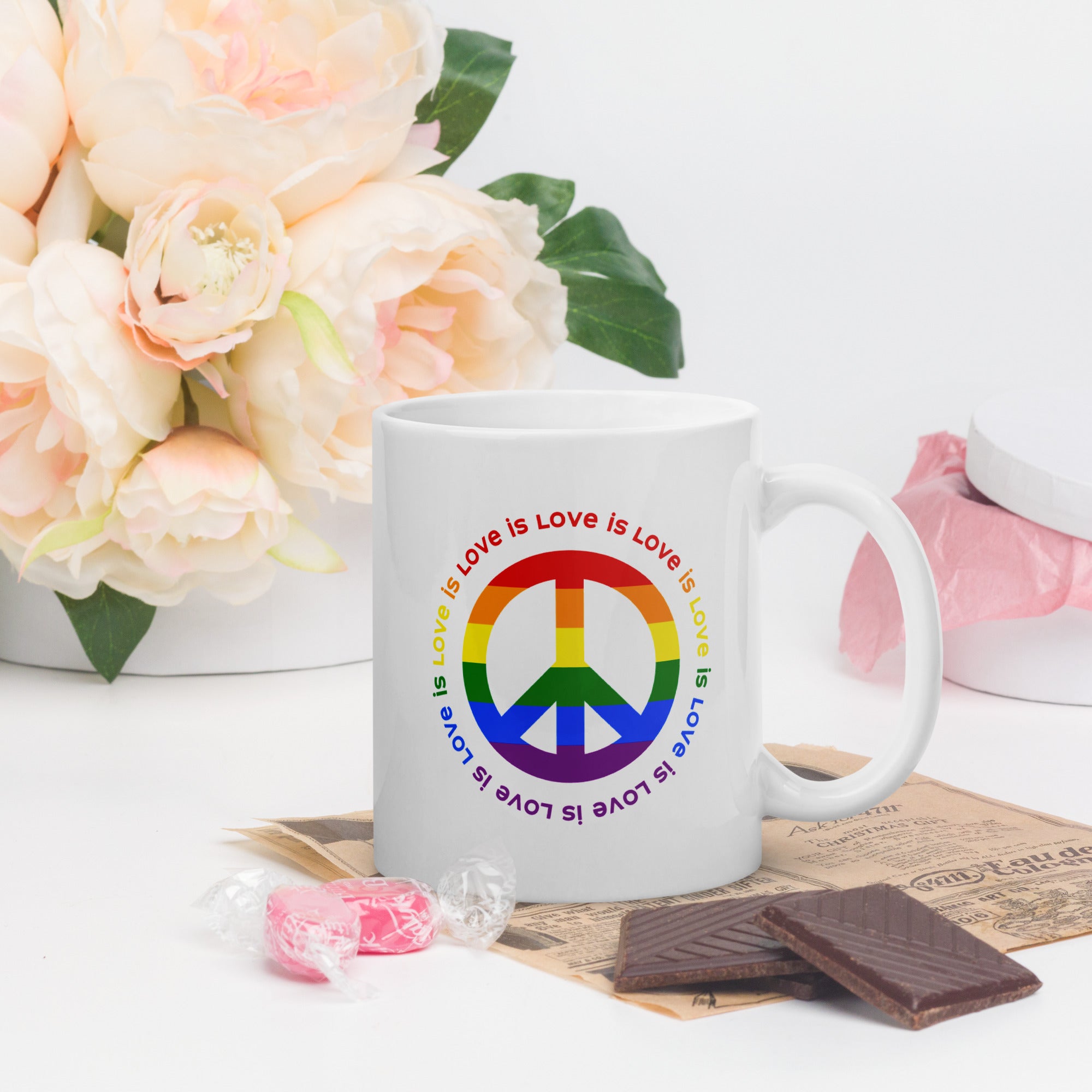 White ceramic coffee mug with a peace sign in the colors of the LGBTQ+ flag, surrounded by repeated "love is love" text. On a tabletop with flowers and candy.
