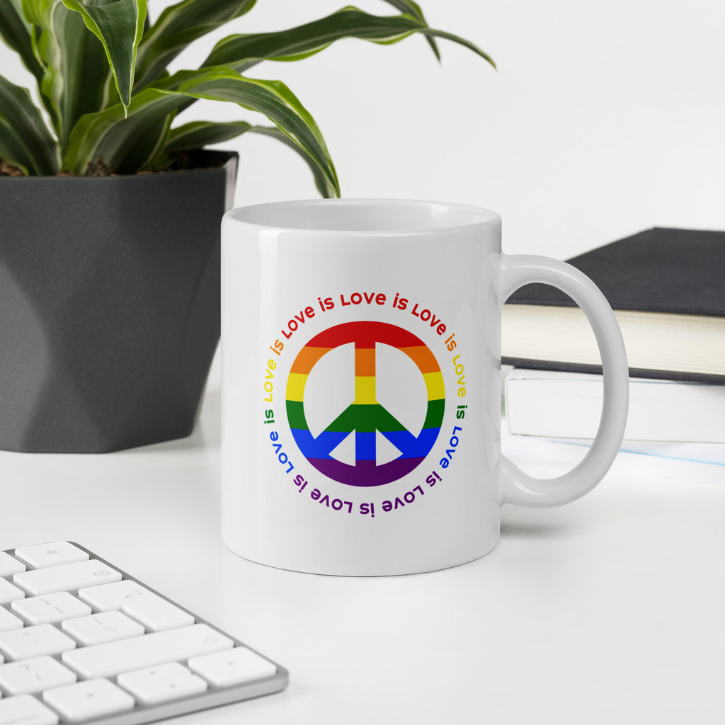 White ceramic coffee mug with a peace sign in the colors of the LGBTQ+ flag, surrounded by repeated "love is love" text. On a desk with a keyboard, plant, and book.