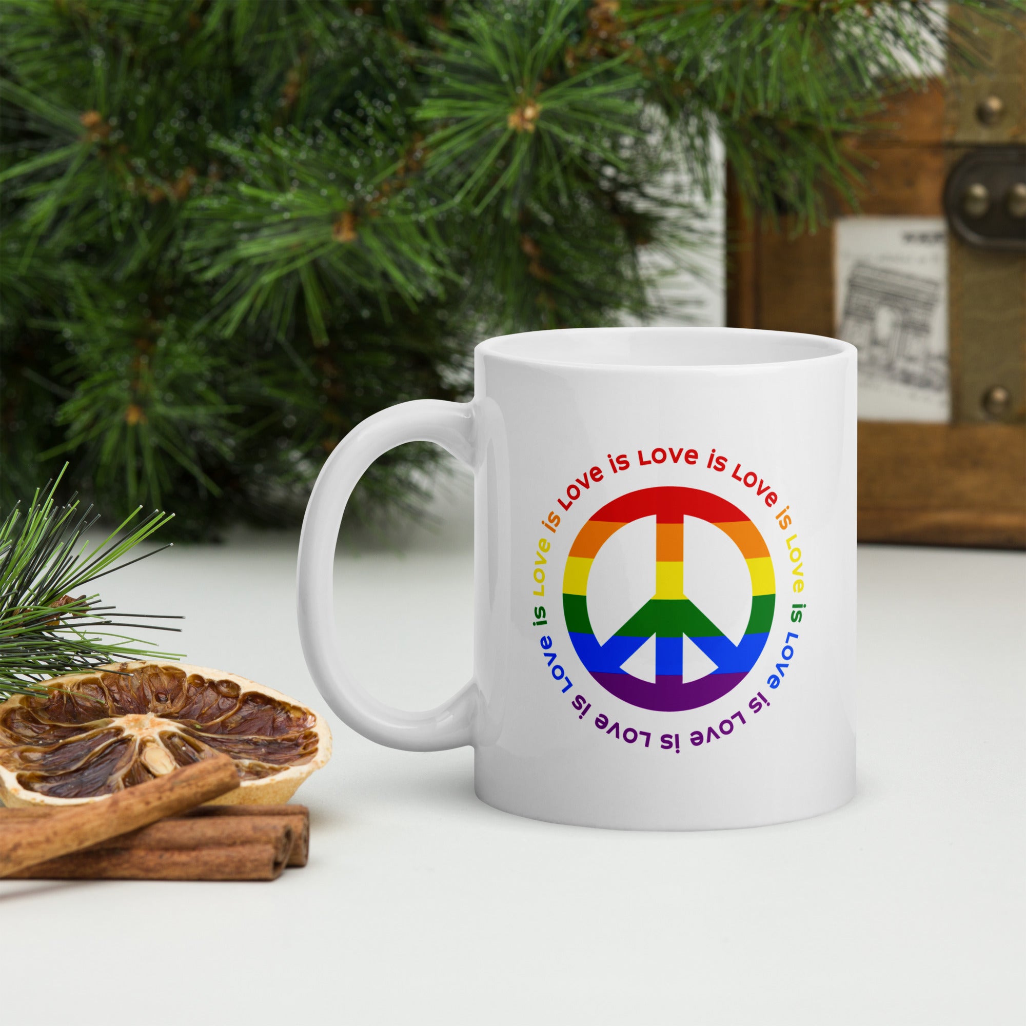 White ceramic coffee mug with a peace sign in the colors of the LGBTQ+ flag, surrounded by repeated "love is love" text. On a tabletop with forest greenery.