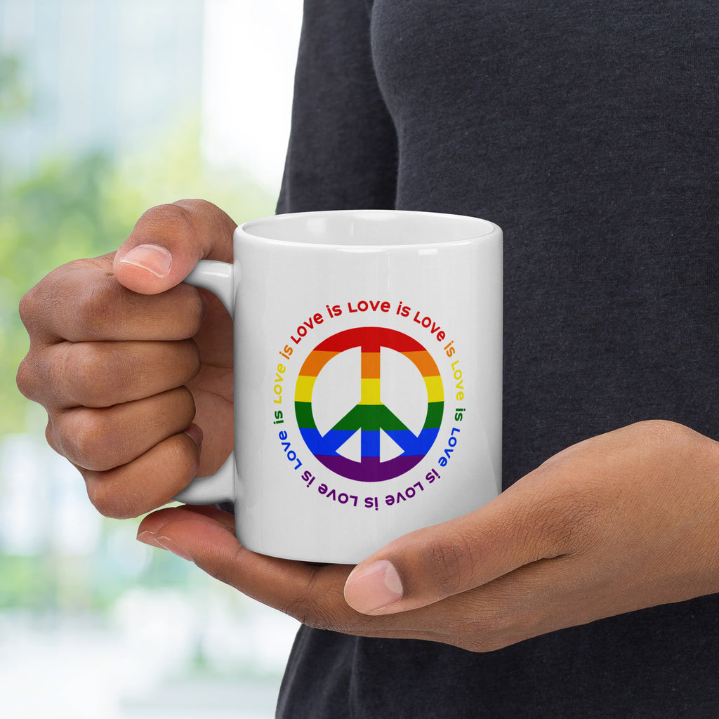 Person holding ceramic coffee mug with a peace sign in the colors of the LGBTQ+ flag, surrounded by repeated "love is love" text. Handle is on the left.