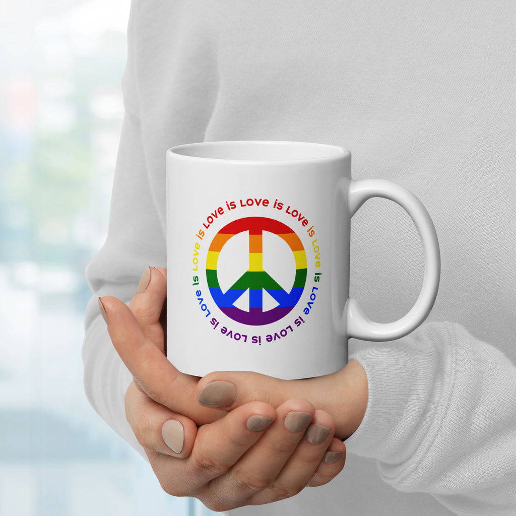 Person holding ceramic coffee mug with a peace sign in the colors of the LGBTQ+ flag, surrounded by repeated "love is love" text. Handle is on the right.