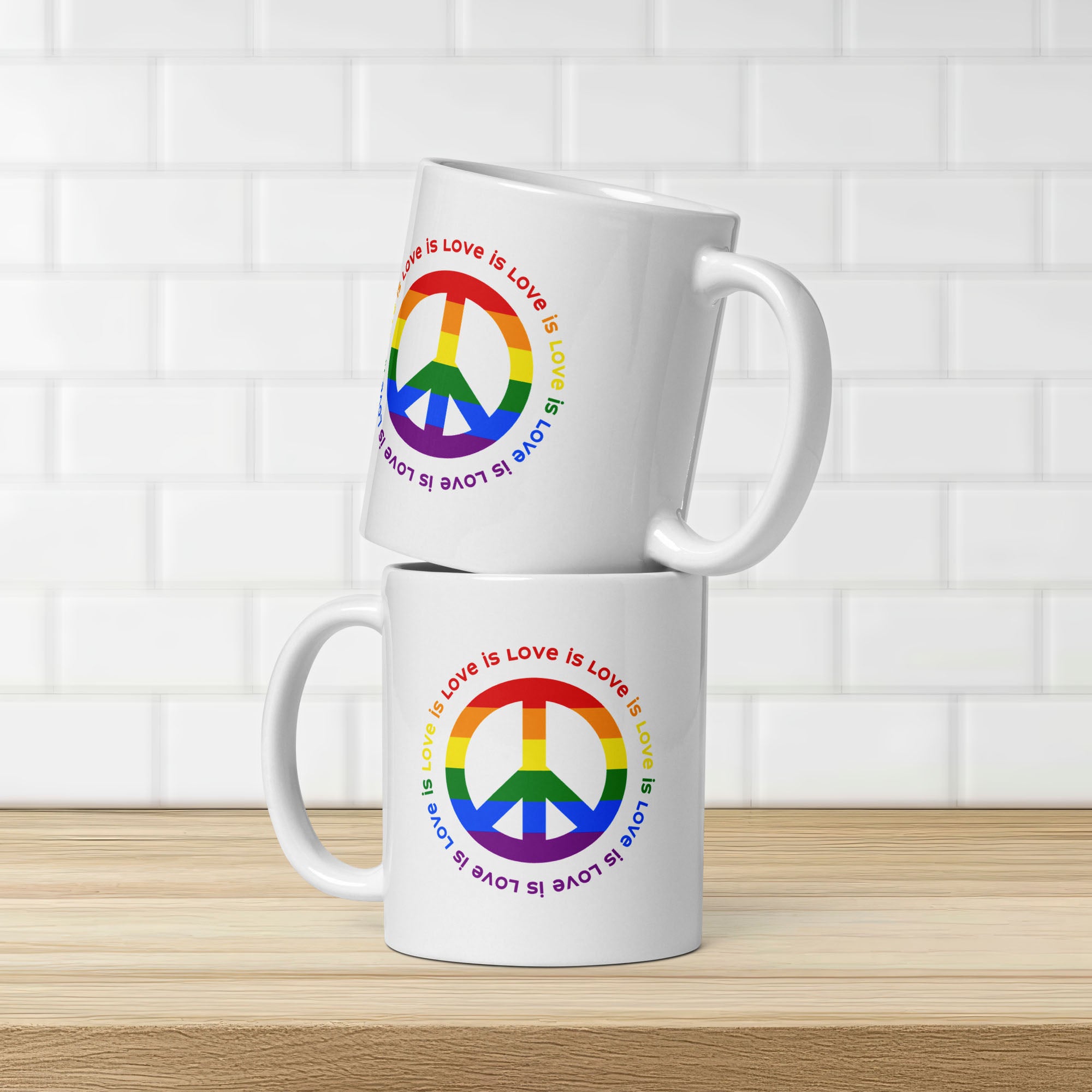 Two ceramic coffee mugs with a peace sign in the colors of the LGBTQ+ flag, surrounded by repeated "love is love" text. Stacked on a kitchen counter with subway tile on the wall.