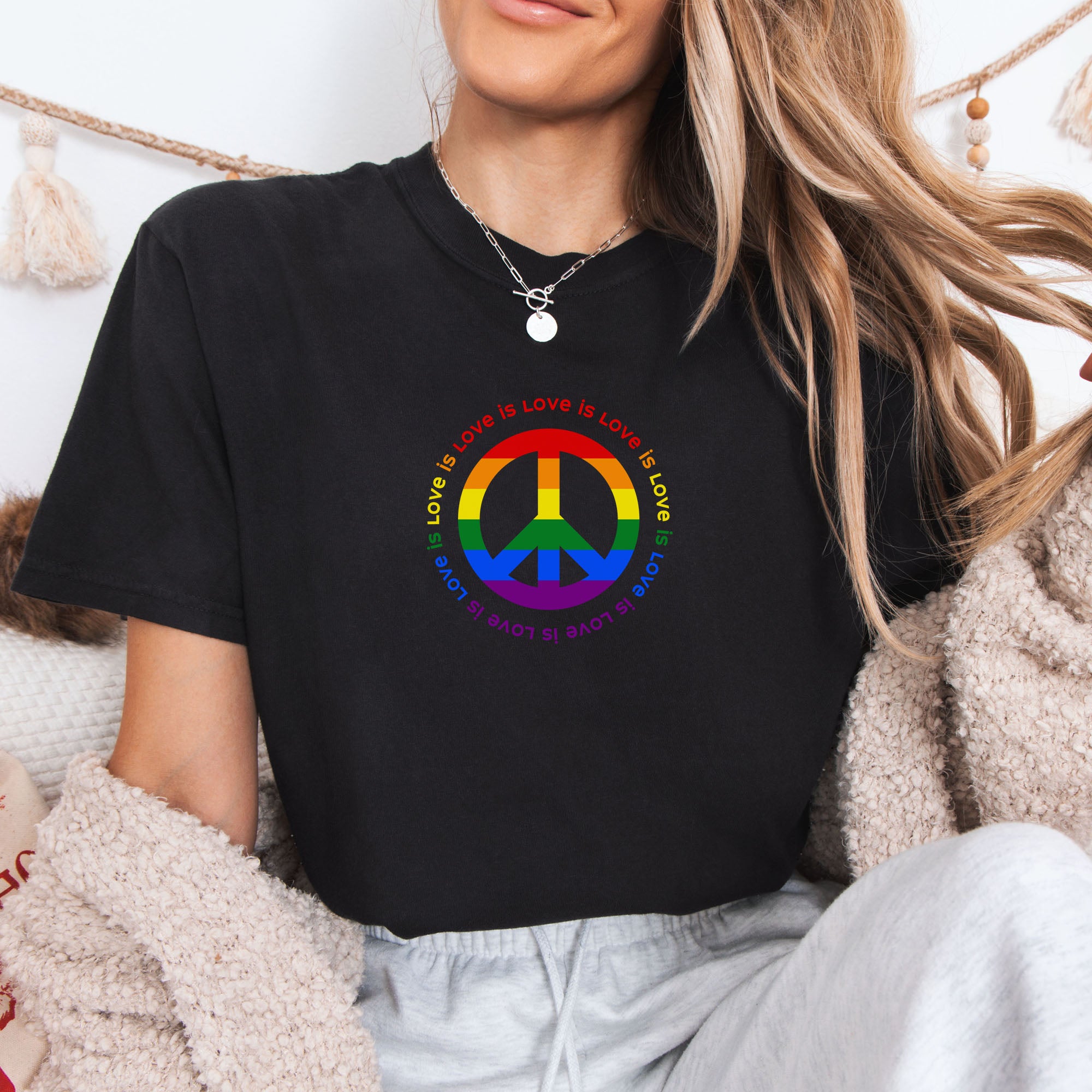 Female model wearing black t-shirt with LGBTQ pride graphic — a peace sign in the LGBTQ flag colors, with the text "love is love" repeated in a circle around the peace sign.