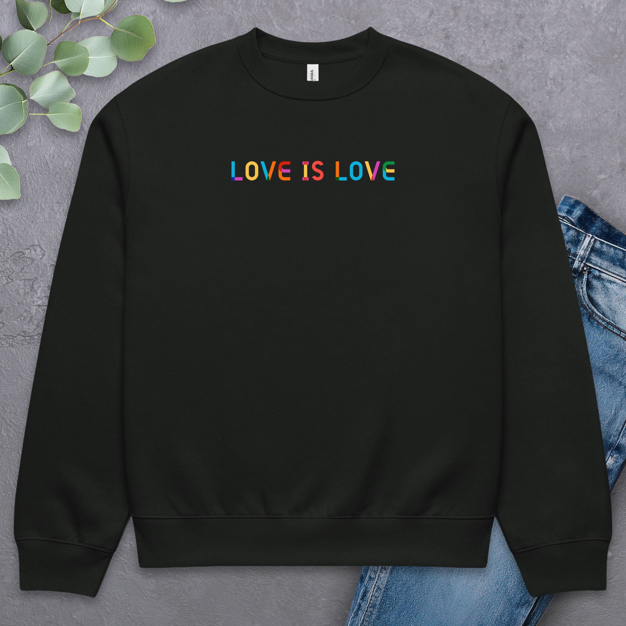 Black Bella+Canvas crewneck sweatshirt with rainbow “love is love” text designed in the Gilbert font, laying flat with a pair of jeans and greenery.