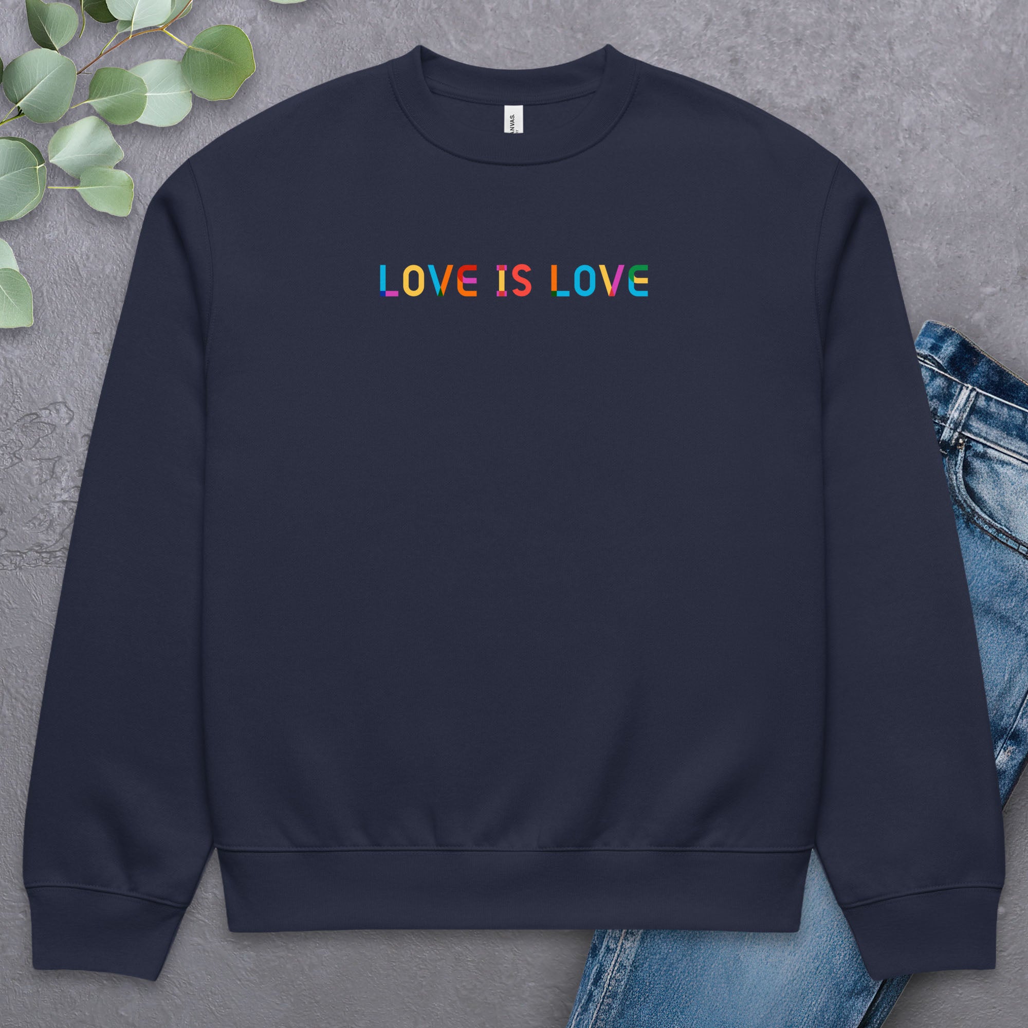 Navy Bella+Canvas crewneck sweatshirt with rainbow “love is love” text designed in the Gilbert font, laying flat with a pair of jeans and greenery.