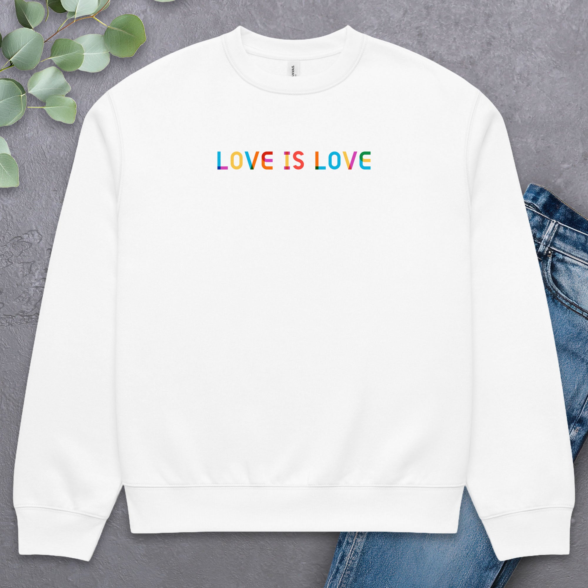 White Bella+Canvas crewneck sweatshirt with rainbow “love is love” text designed in the Gilbert font, laying flat with a pair of jeans and greenery.